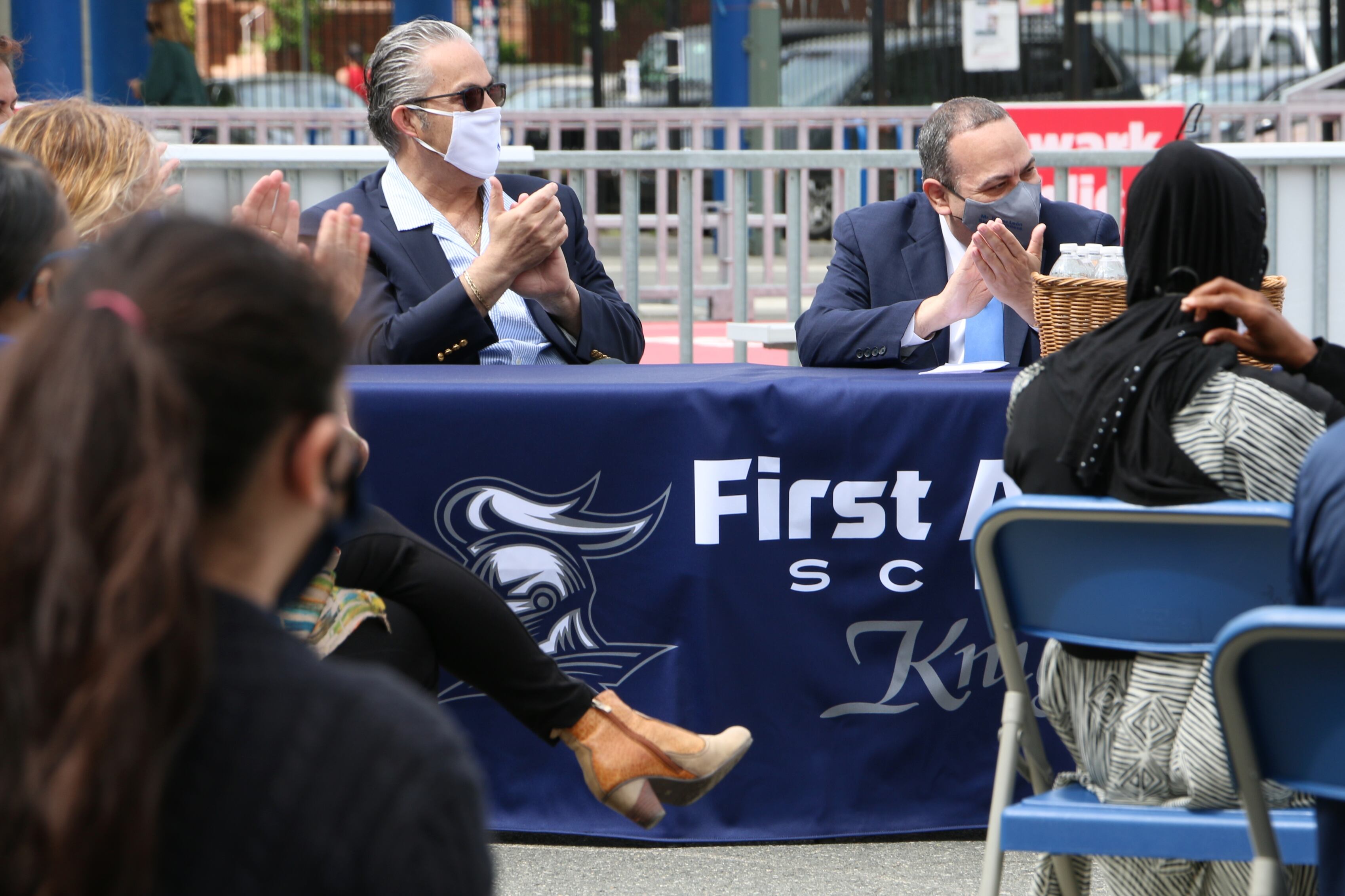 Newark schools Superintendent Roger León sits beside Newark Teachers Union President John Abeigon at a table covered in a blue cloth. Both men are clapping and wearing masks.