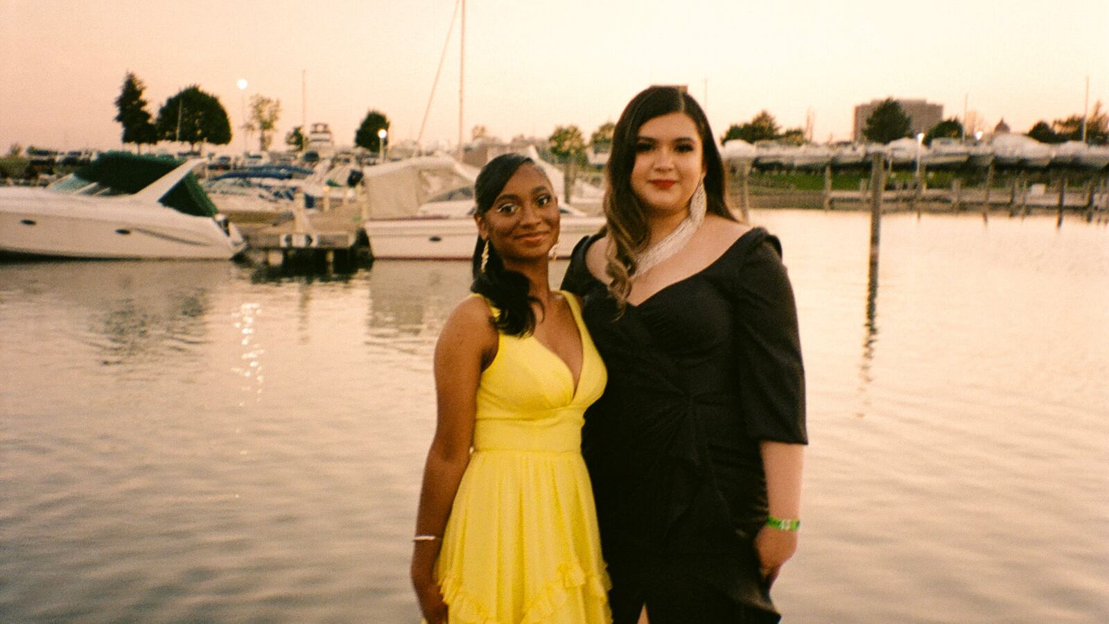 Two young women, one wearing a bright yellow dress and the other wearing a black dress, pose for a portrait together on a dock, boats swaying on the water in the background.