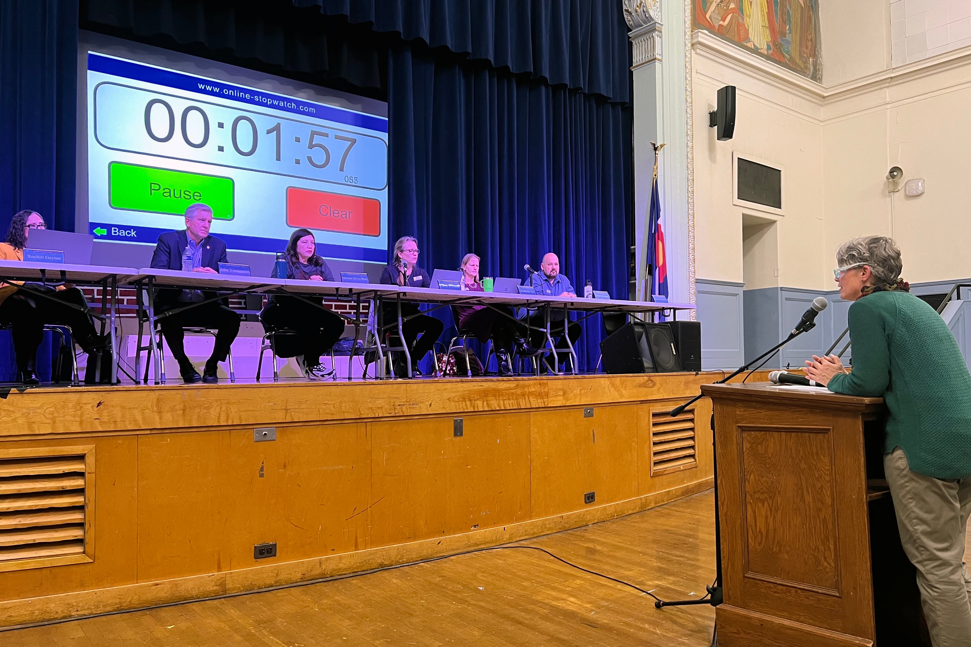 A group of people sit at a long table on stage while one person stands and speaks from behind a wooden podium in a school auditorium.