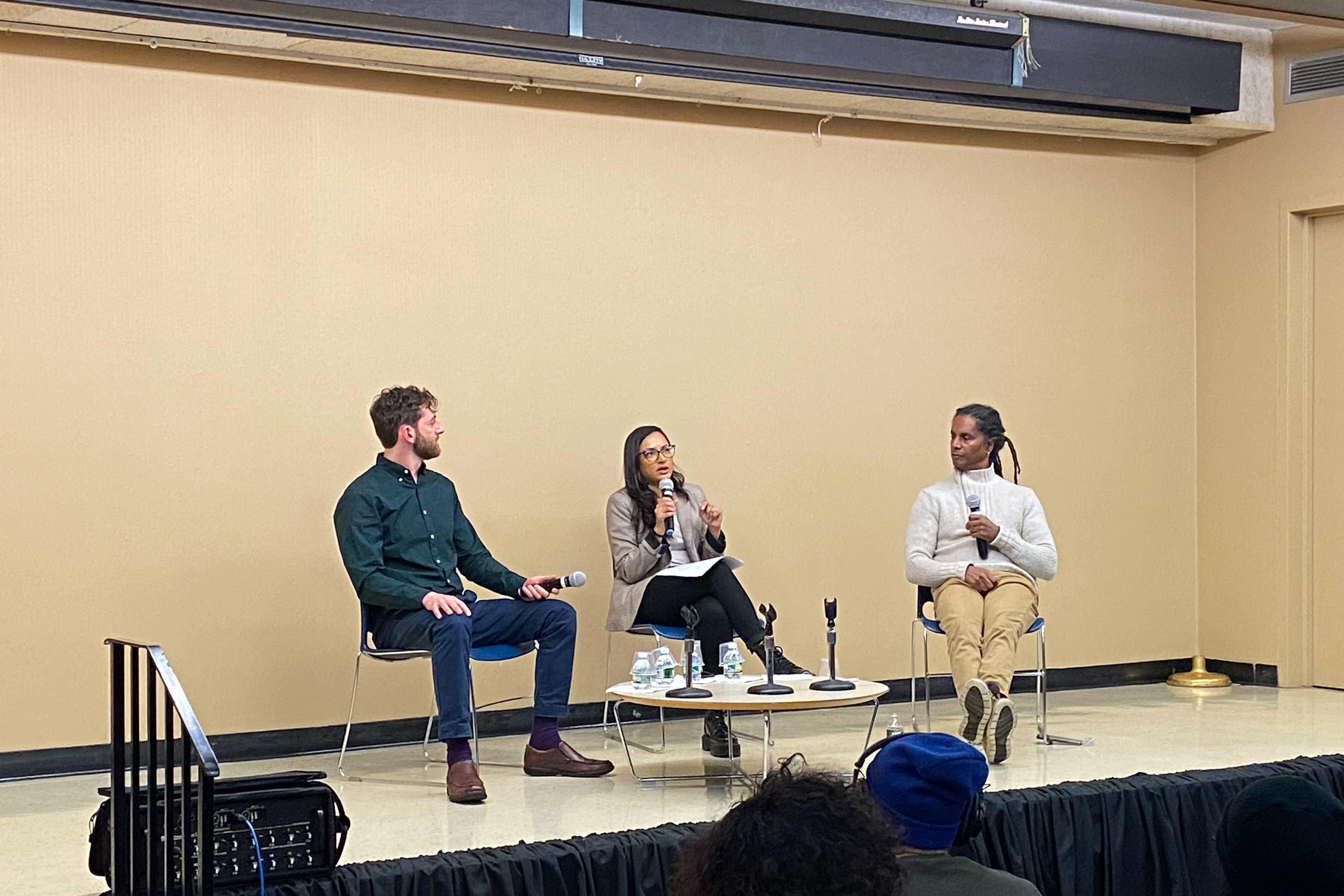 Two men and a woman sit on stage with microphones during a live podcast taping.