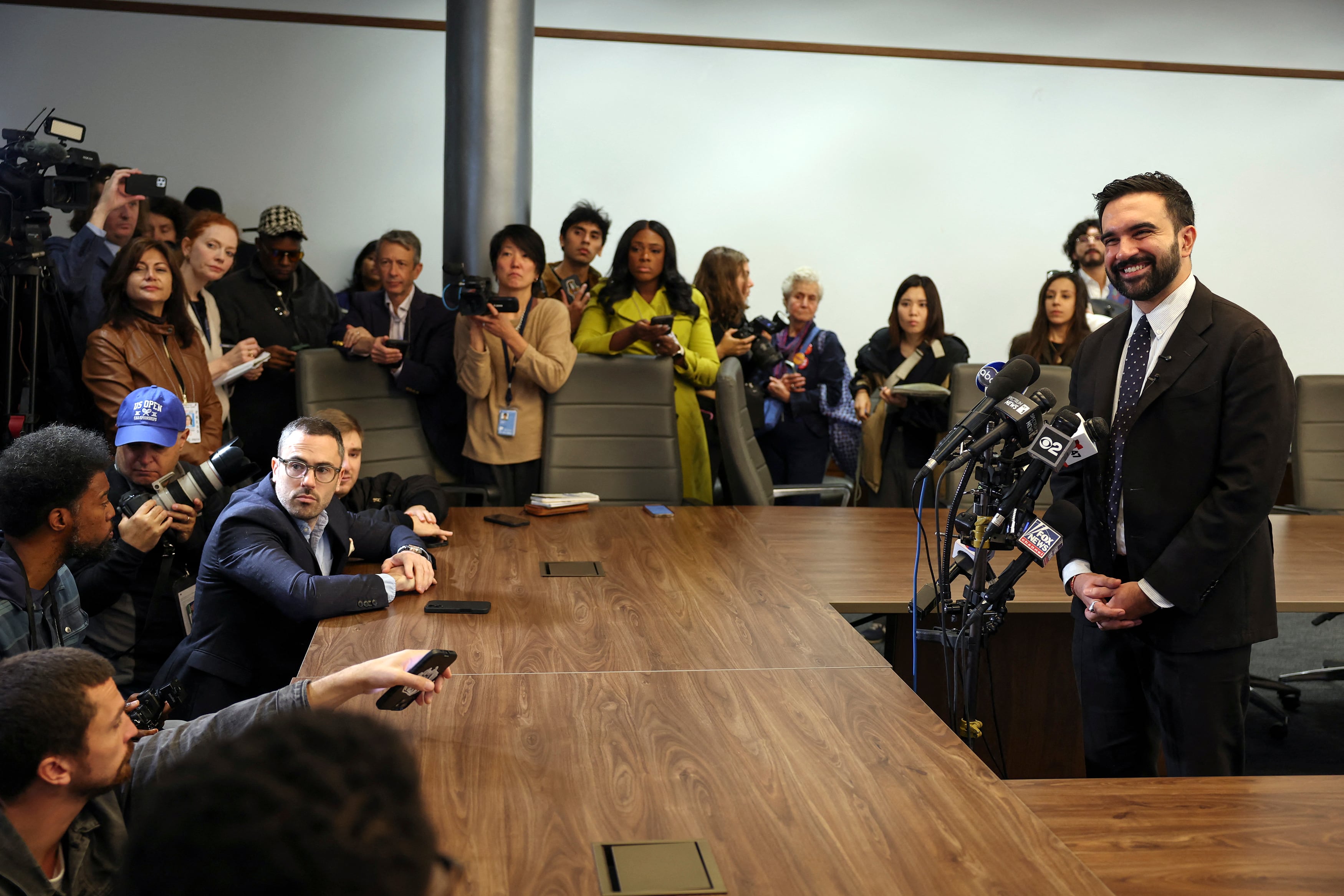 A photograph of a man in a suit standing behind a group of wooden tables speaking into a dozen microphones and in front of a large group of reporters and people.