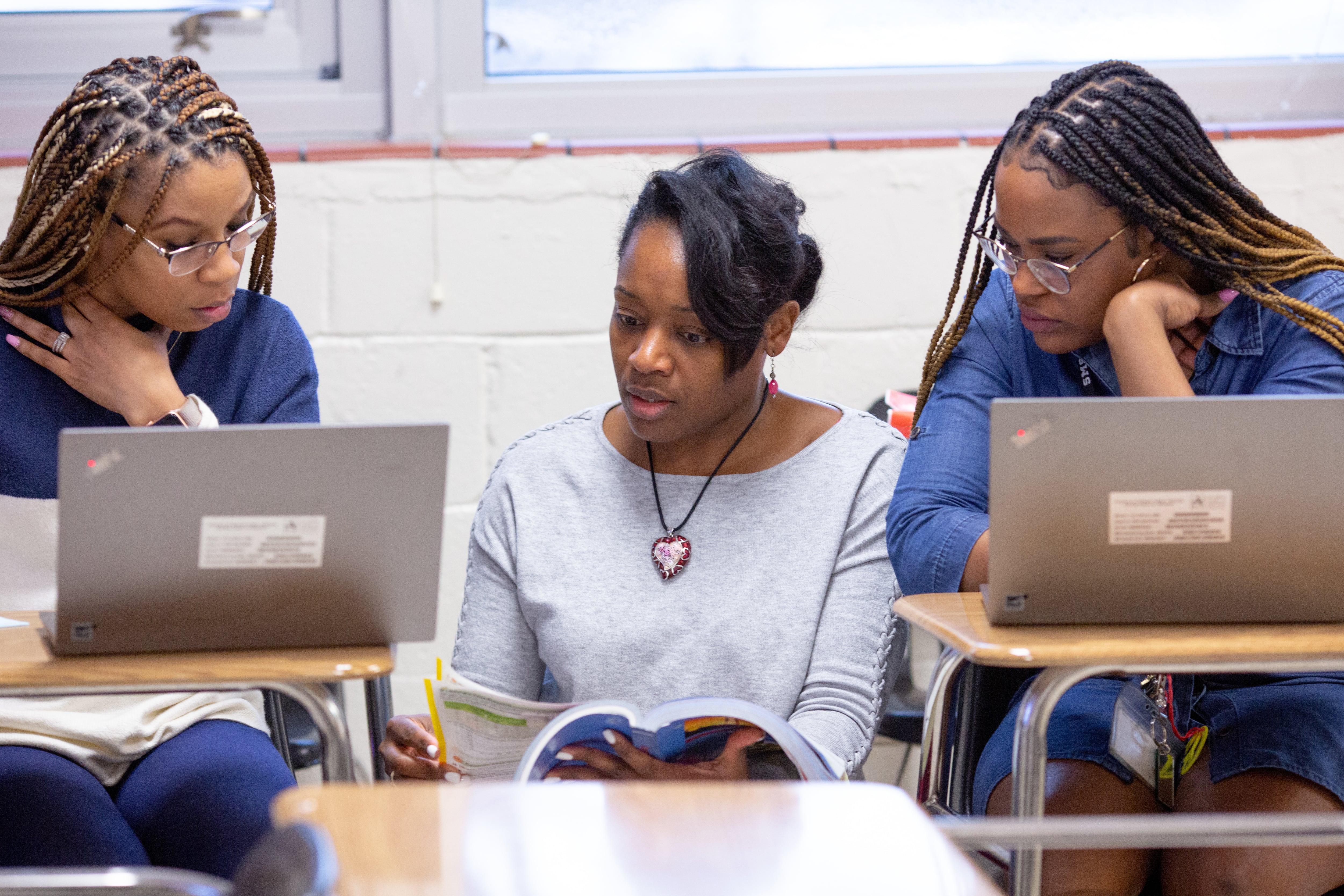 Two young women sit at desks in a classroom with laptops open, looking to a teacher who sits between them going through a textbook.