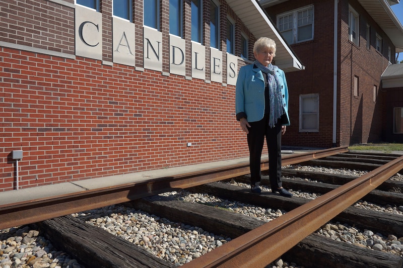 A woman with short white hair and wearing a bright blue jacket stands outside on a railroad track in front of a red brick building that has the word "Candles" on the side.