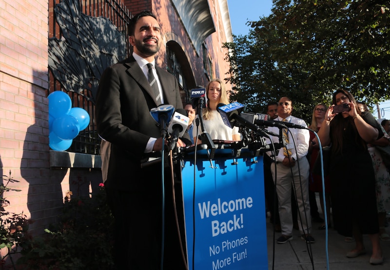 A photograph of a man in a suit standing from behind a podium with a group of people outside.