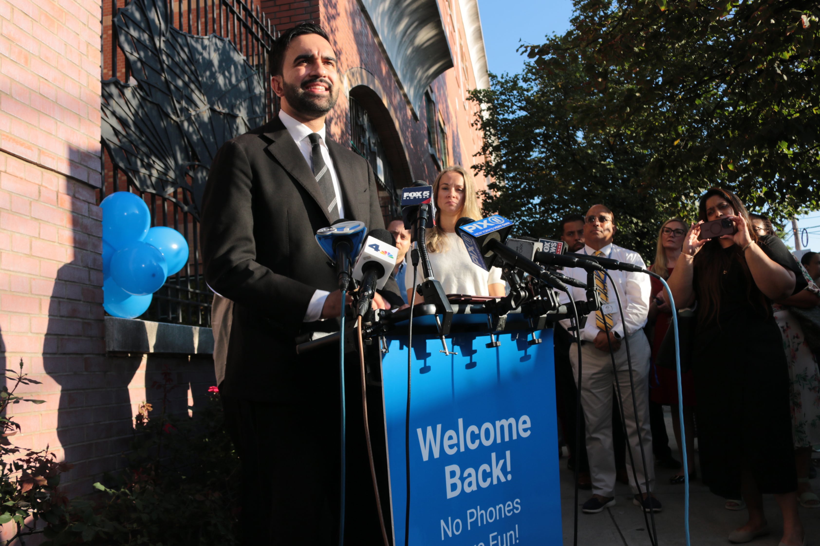 A photograph of a man in a suit standing from behind a podium with a group of people outside.