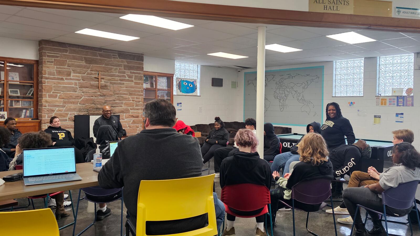A group of adults and high school students gather around in a wonky circle in a room with a large brick wall in the back.