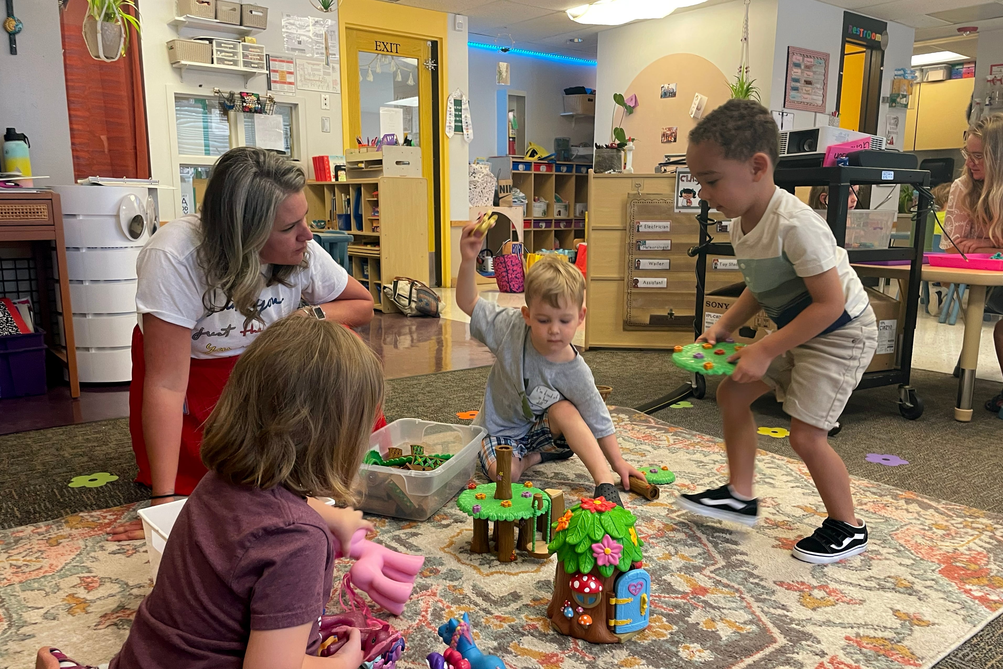 Three preschool children play with green and brown plastic tree houses while a teacher looks on.