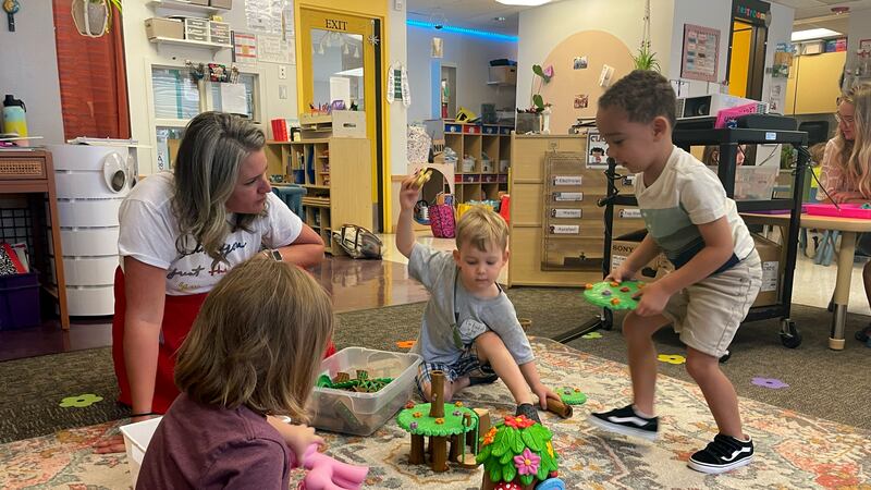 Three preschool children play with green and brown plastic tree houses while a teacher looks on.
