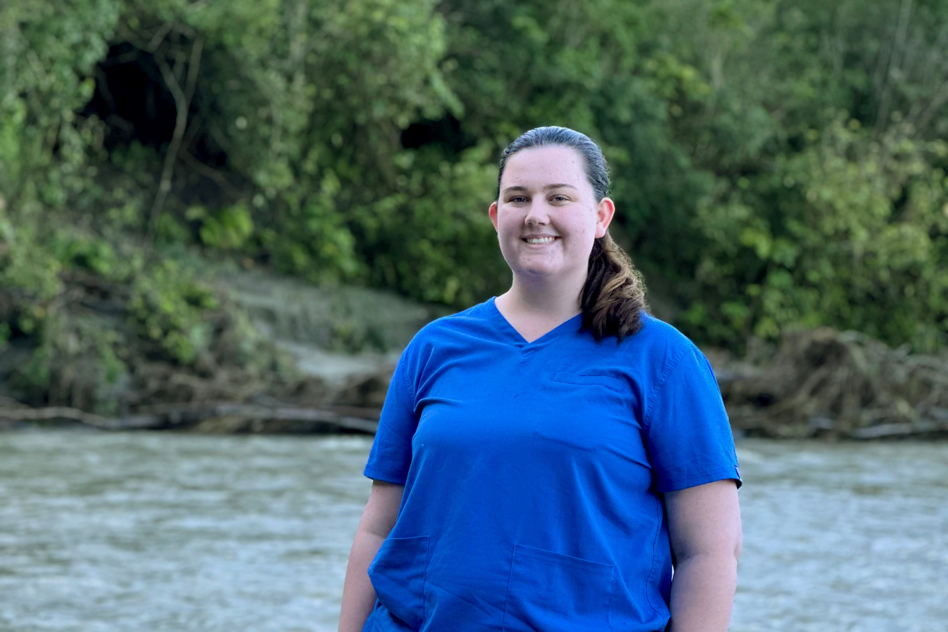 A woman wearing blue scrubs stands in front of a body of water and green trees.