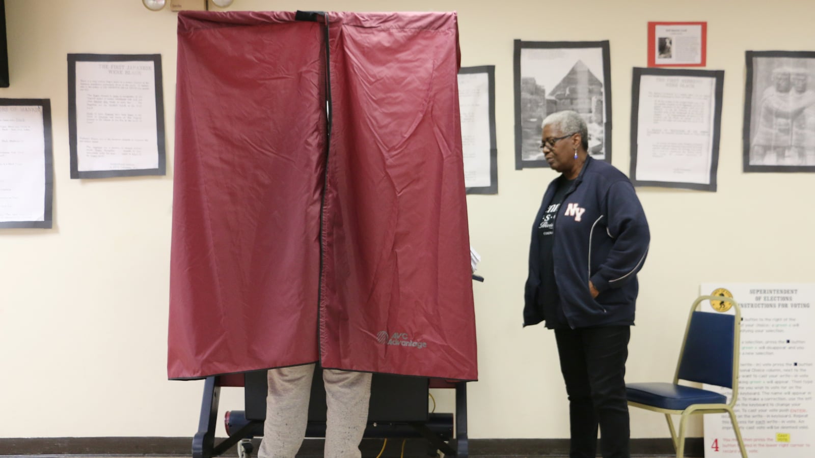 One person stands outside of a voting booth while one person is using the voting booth in a room with large picture frames on the wall in the background.