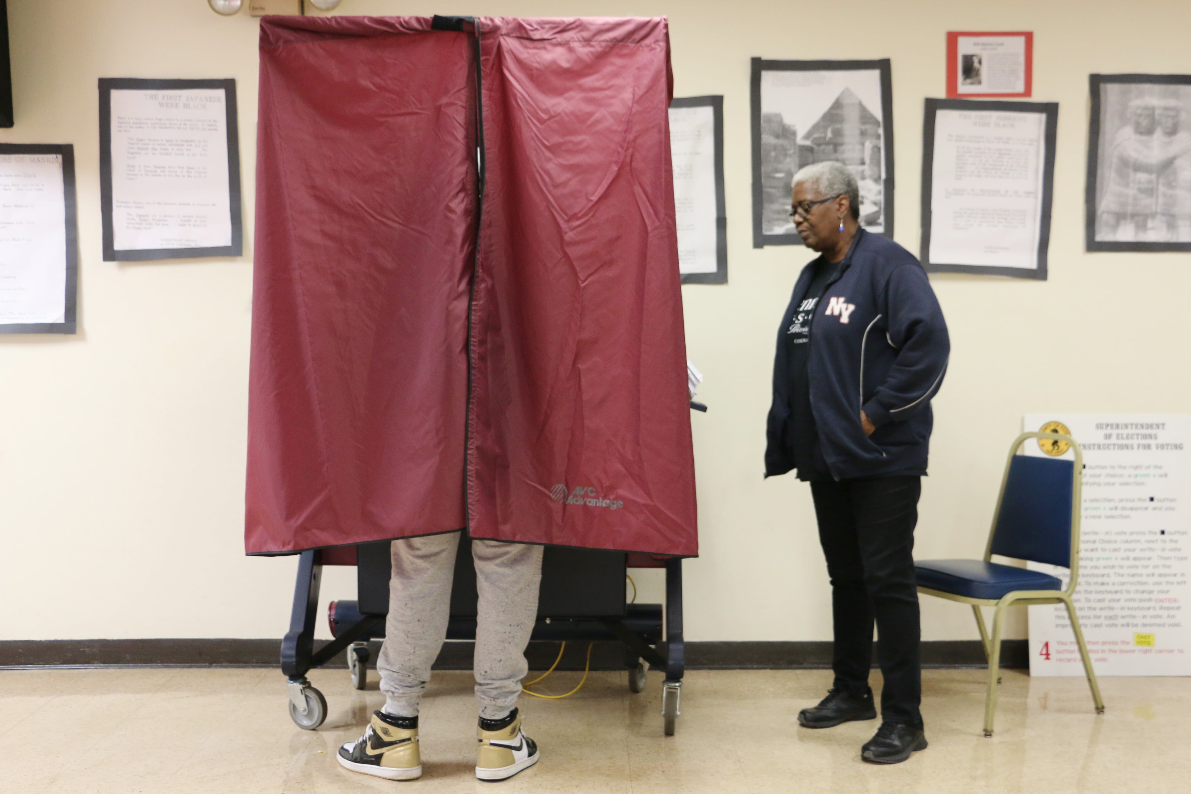 One person stands outside of a voting booth while one person is using the voting booth in a room with large picture frames on the wall in the background.