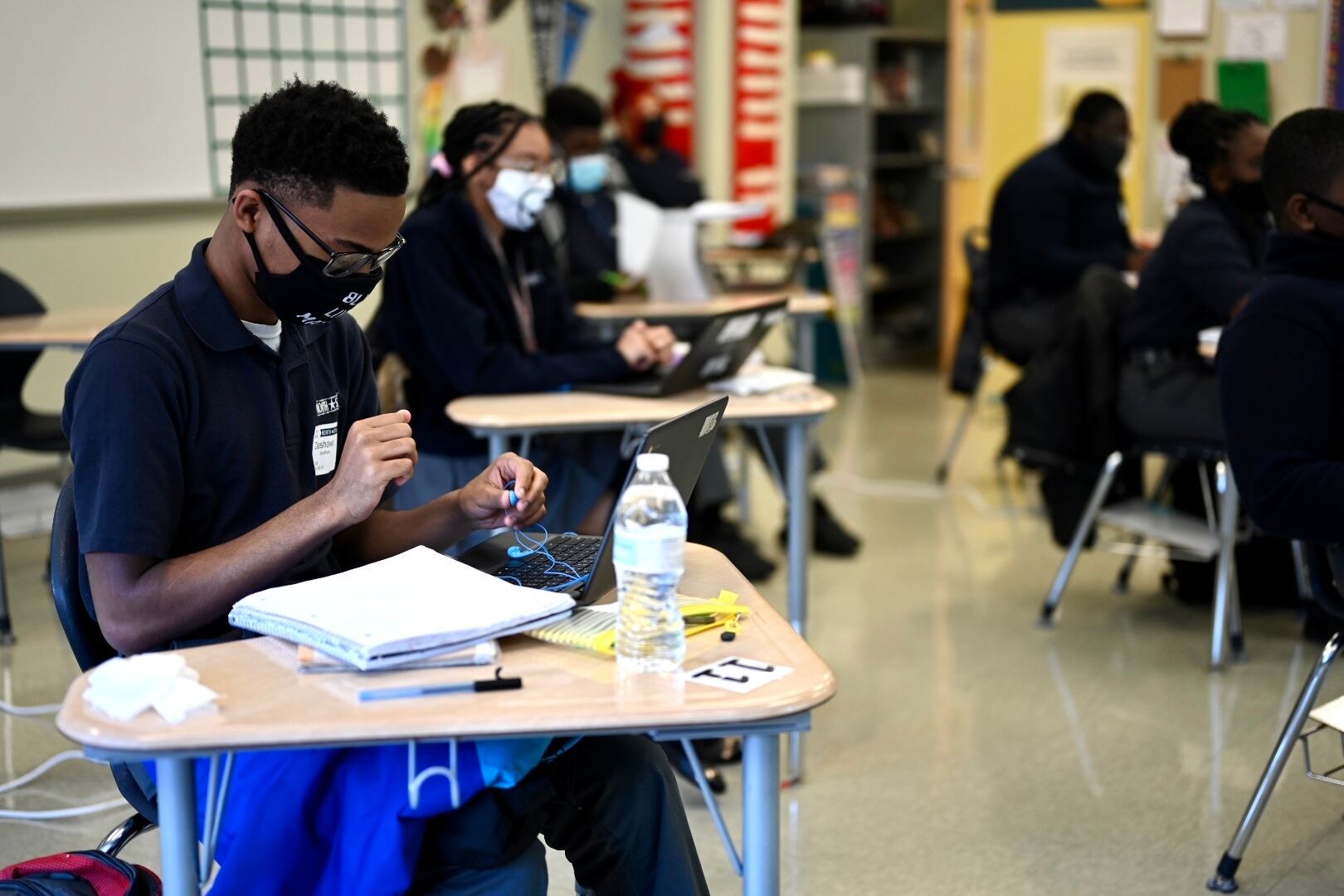 A young man works at his desk, wearing glasses, a protective mask, and a blue polo shirt. Several other students work at their desks around him in a classroom.
