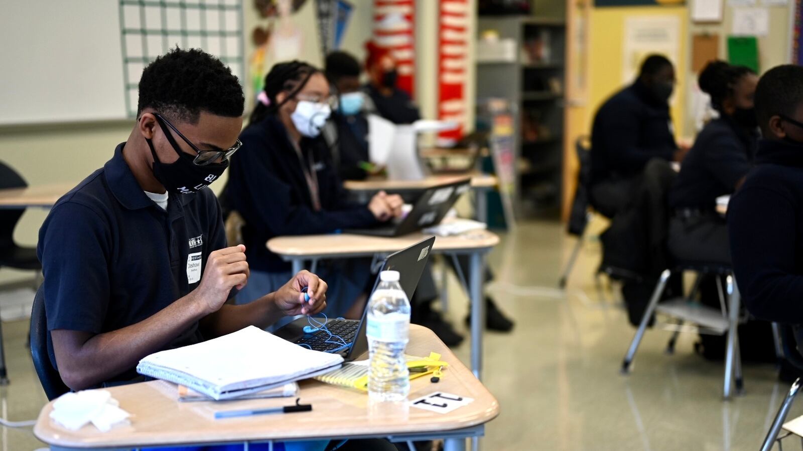 A young man works at his desk, wearing glasses, a protective mask, and a blue polo shirt. Several other students work at their desks around him in a classroom.