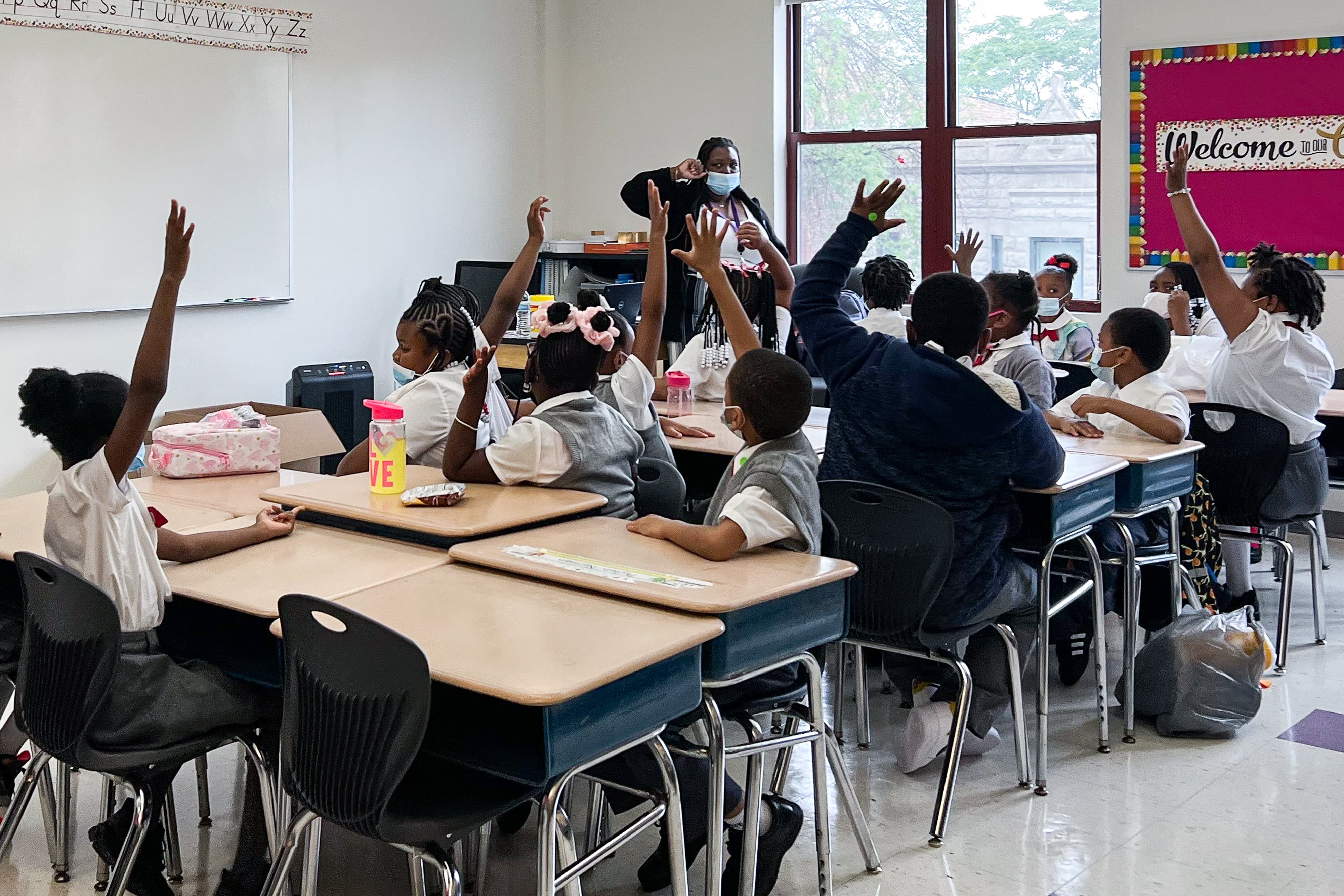Uniformed students in second grade sitting at desks raise their hands as their teachers looks at them in a classroom. The students and teacher are all wearing masks.
