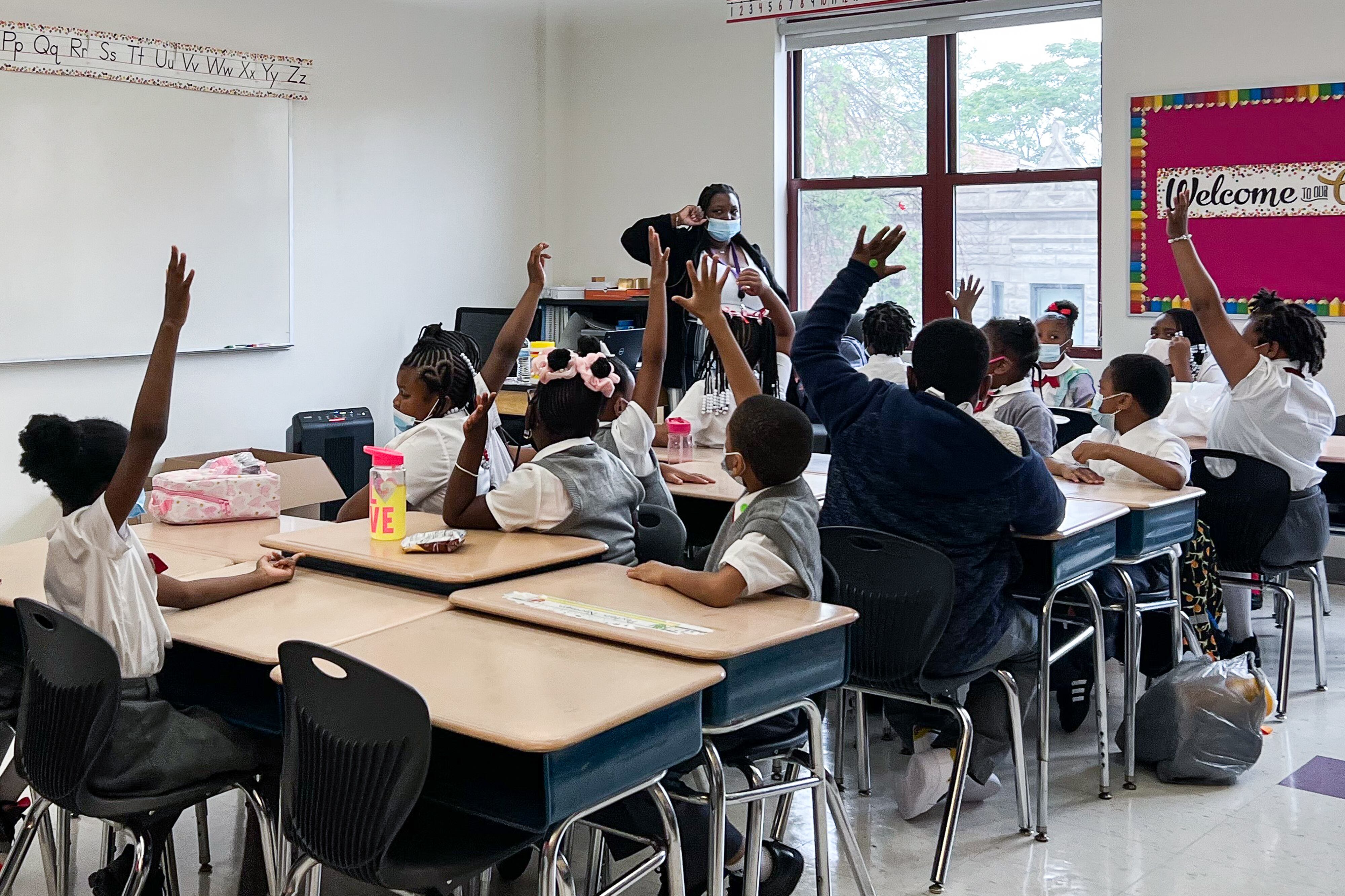 Uniformed students in second grade sitting at desks raise their hands as their teachers looks at them in a classroom. The students and teacher are all wearing masks.