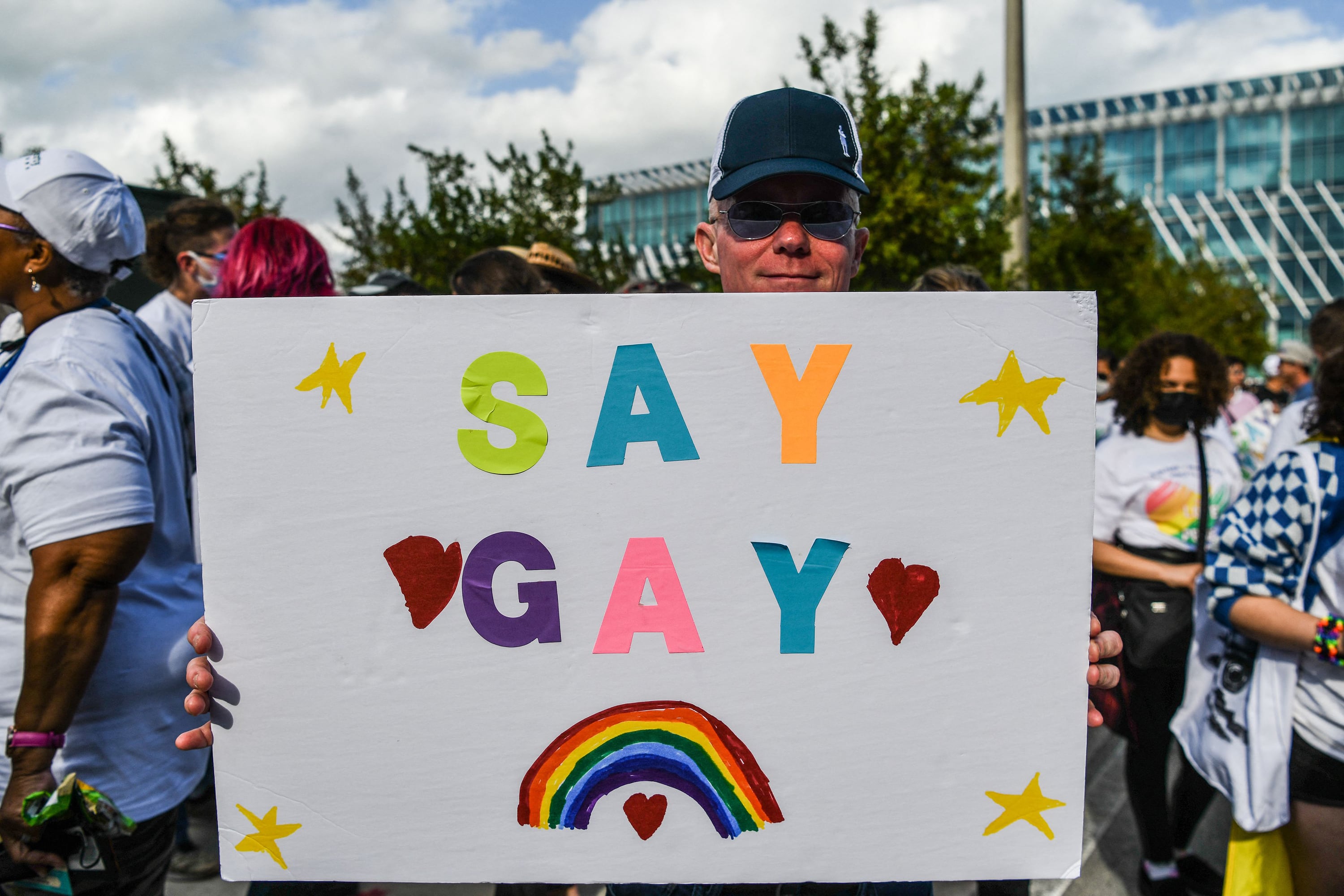 A person wearing a hat and holding a giant white sign with colorful letters that say "SAY GAY" with a rainbow, stars and hearts with people standing in the background.