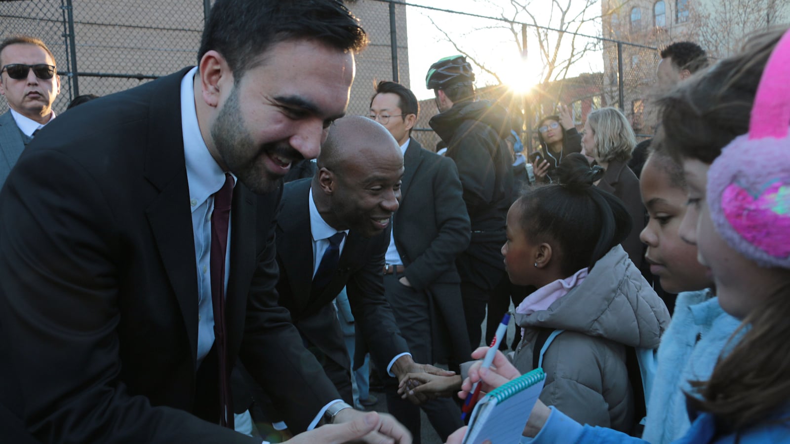 A photograph of two men in suits greet young children outside on a cold day.