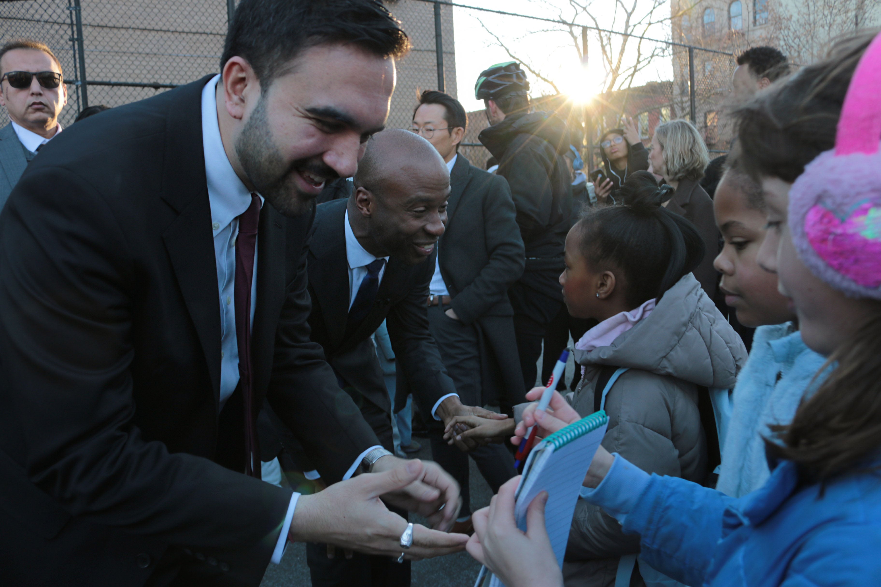 A photograph of two men in suits greet young children outside on a cold day.