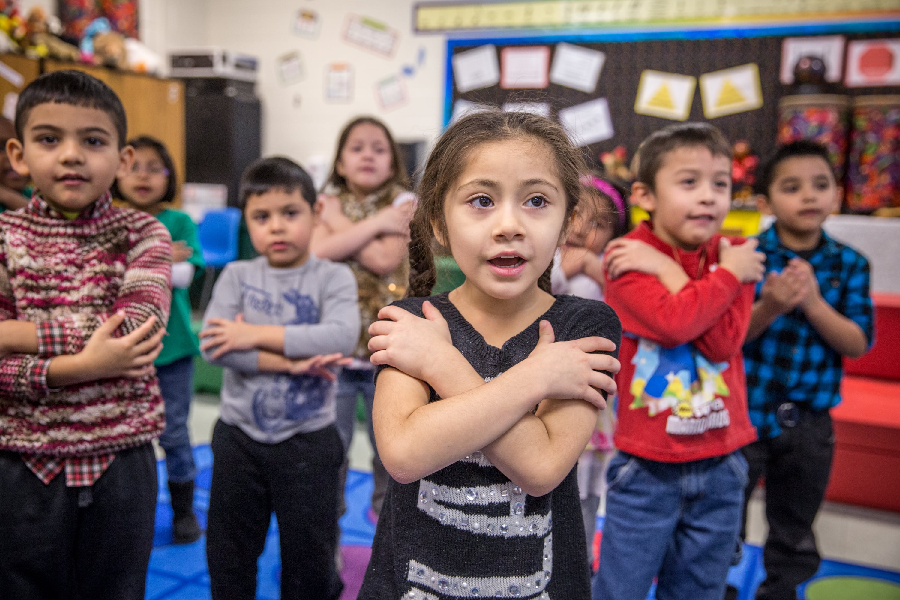 A group of young children touch their shoulders with opposite hands during a classroom activity.