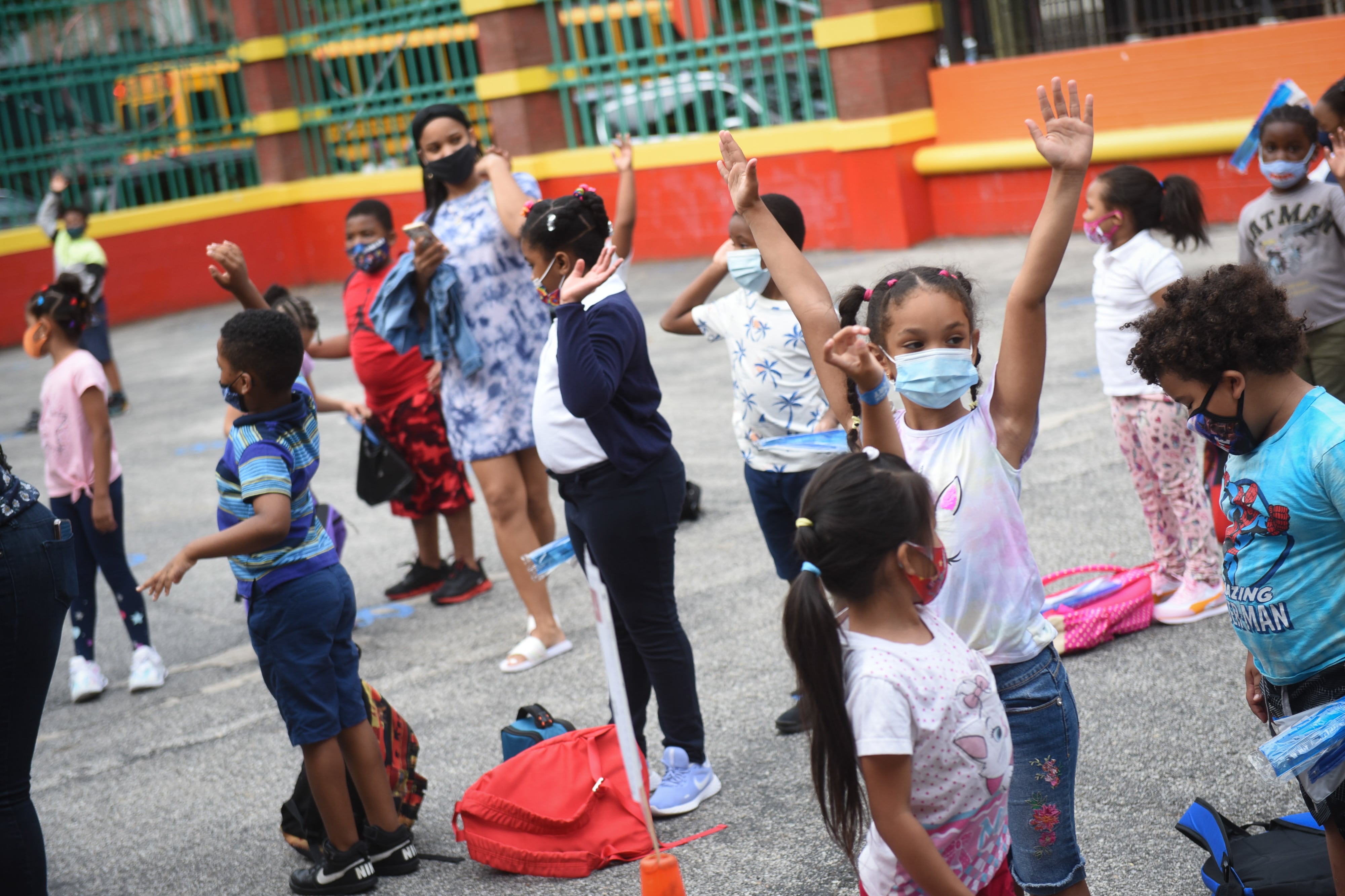 About a dozen elementary-age students, some with their hands raised, and an adult stand on a playground with a wall and fence in the background.
