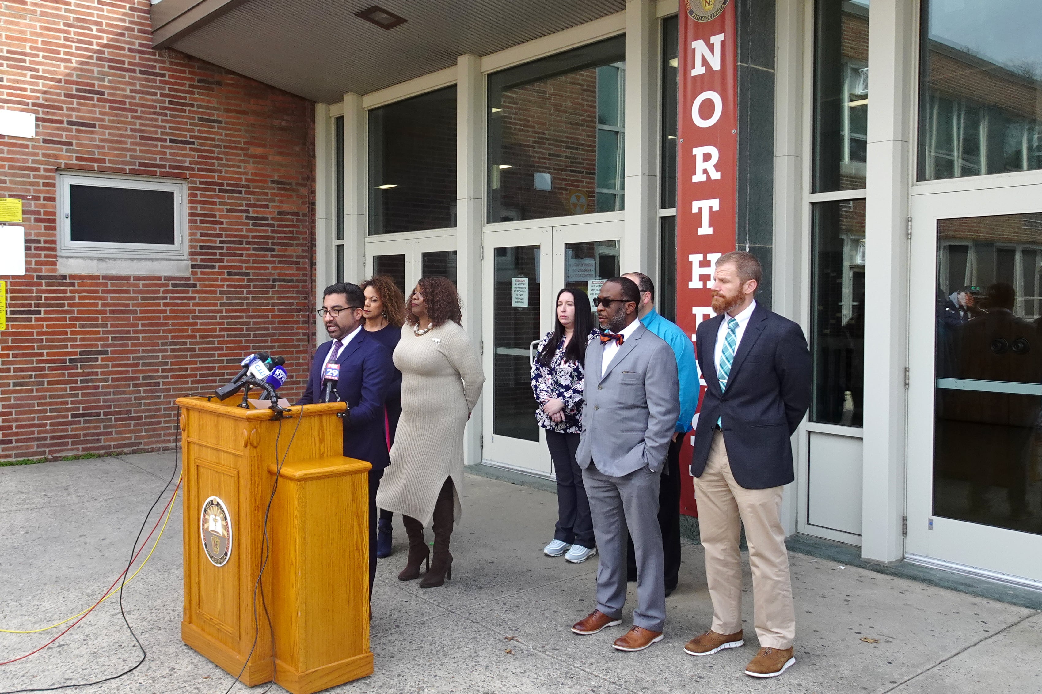 A group of people stand behind a podium in front a glass doors to the front entrance of a building.