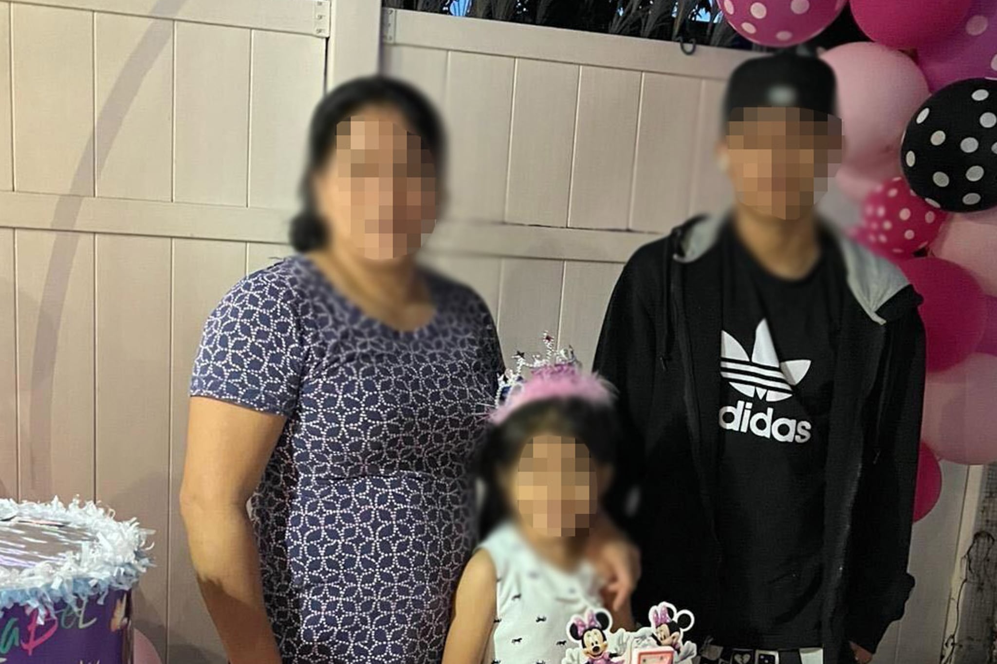 A woman, a man and a 5-year-old child stand behind a table with a pink birthday cake on it. Their faces are blurred out.
