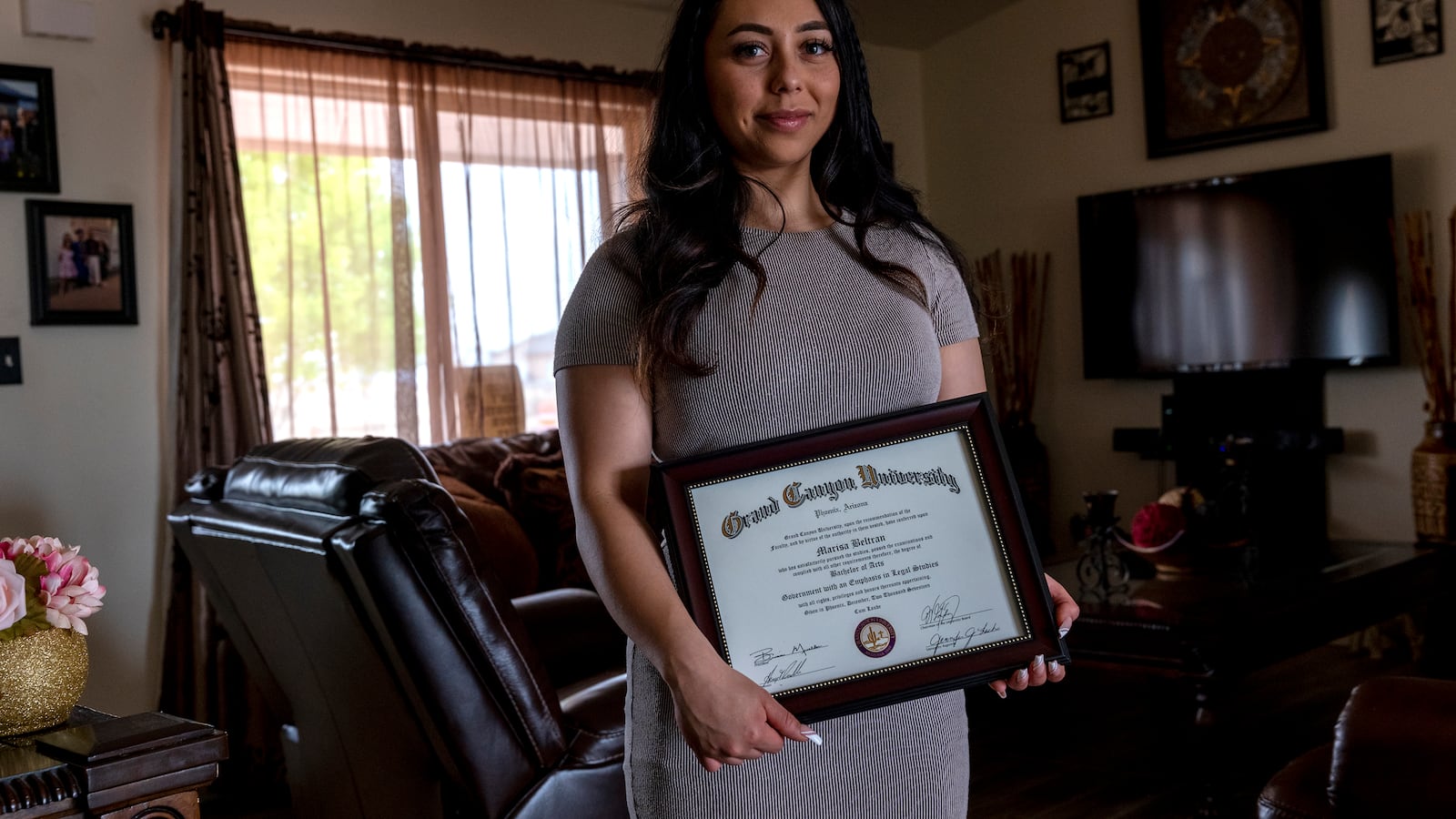 A young woman stands in a living room holding her college diploma from Grand Canyon University.