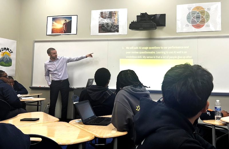 A man in a light shirt points at a board in front of students seated at tables.