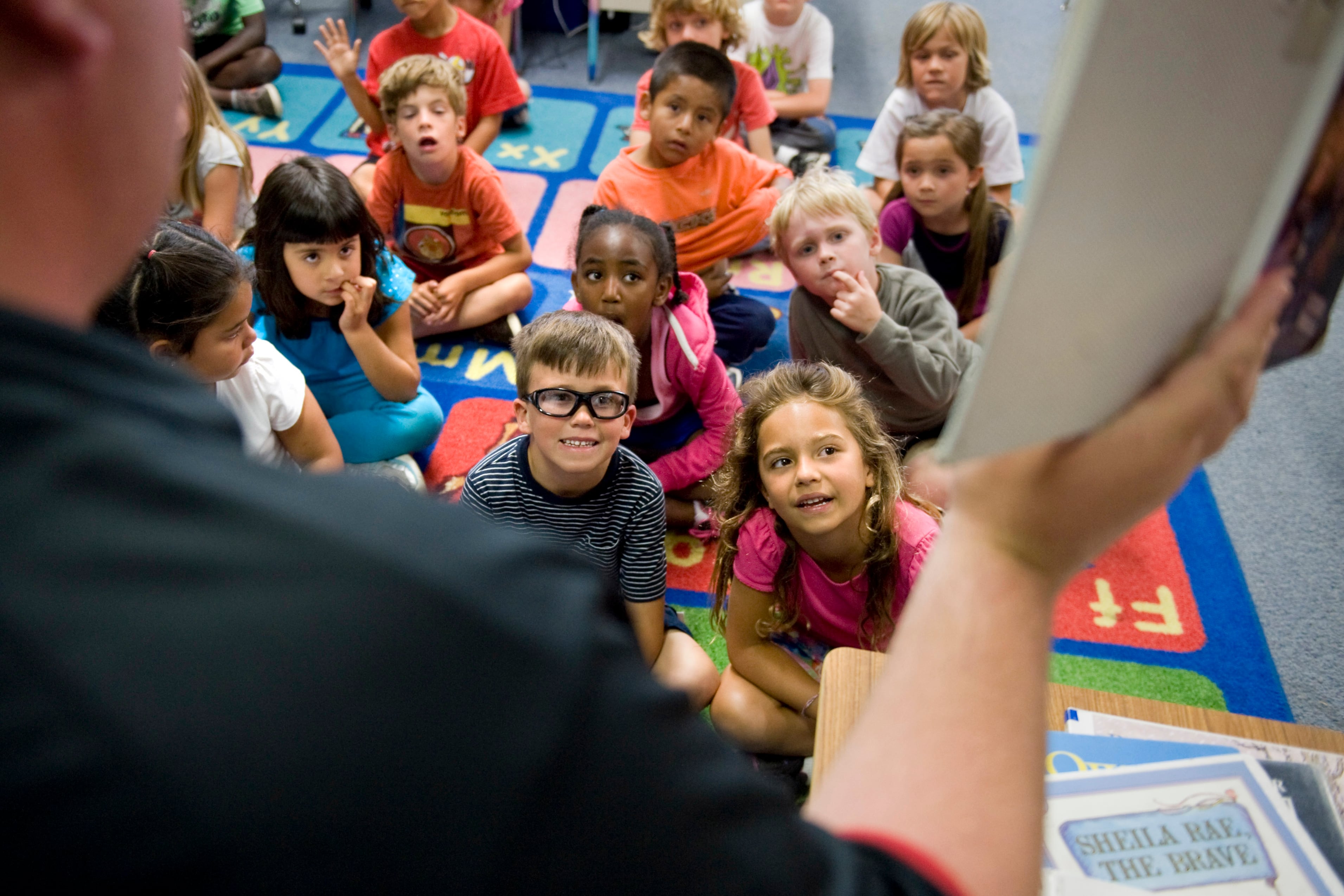 Young students sit on a carpet looking at a teacher holding part of a lesson