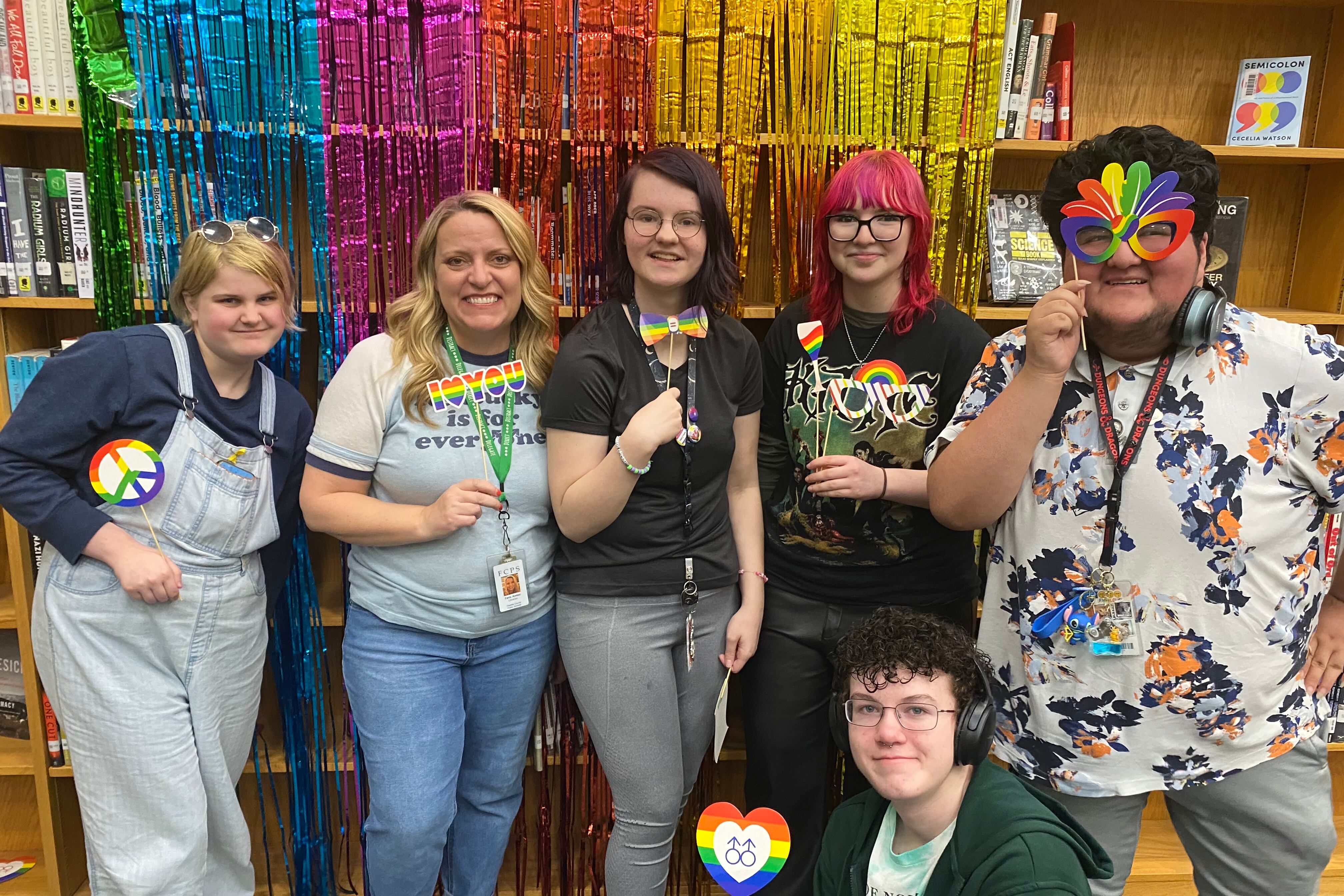 A group of students and a teacher is standing together in front of rainbow ribbons. They are holding rainbow sunglasses and other photo props.