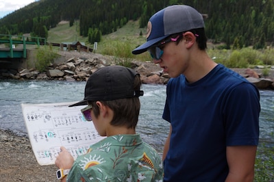 Two teenage boys, one much taller than the other, study a chart showing different types of insects. They are standing next to a river in the mountains. A bridge is visible in the background.