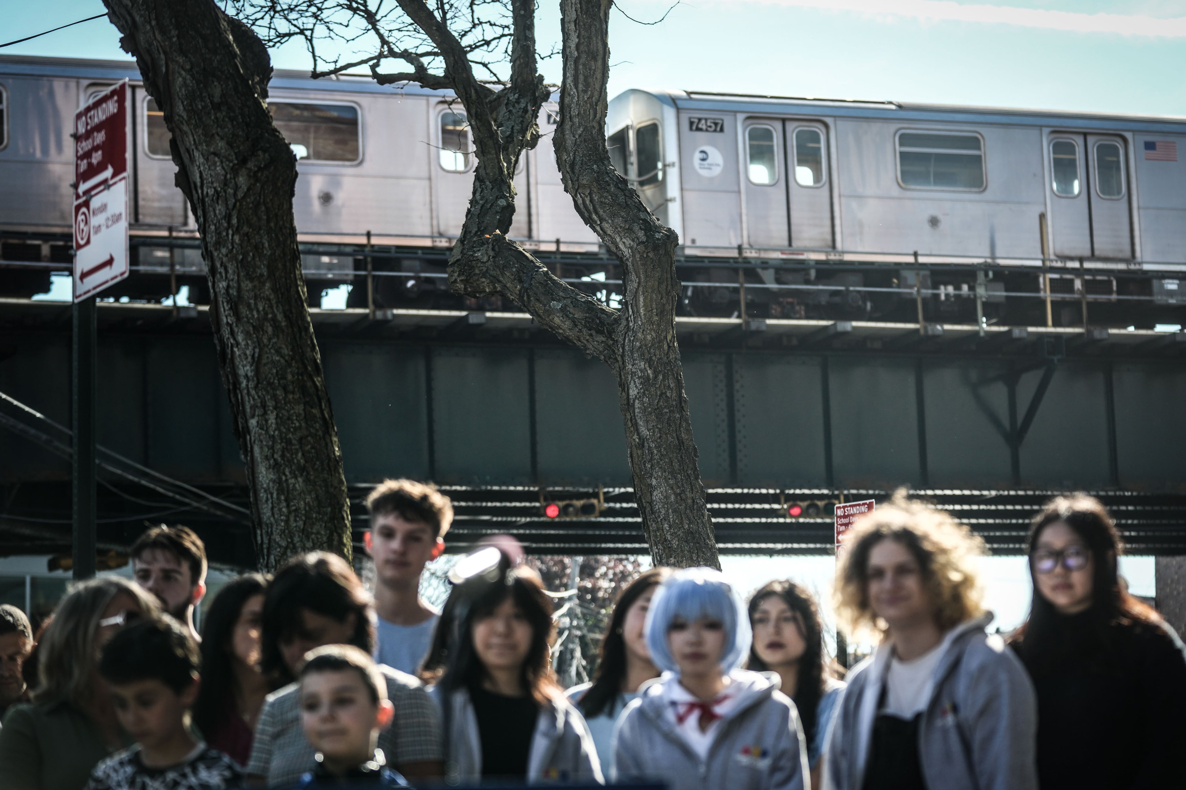 A group of people stand in the foreground while a subway train passes overhead in the background.
