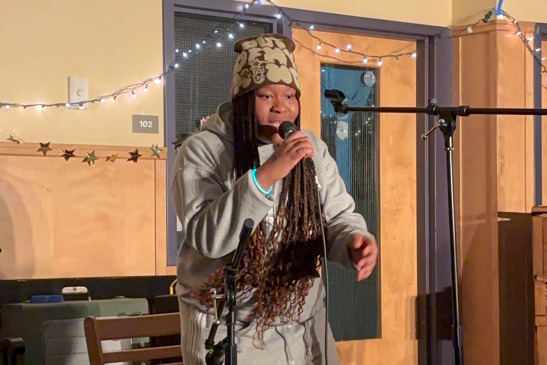 A Black high school senior with long braids performs in a school building holding a microphone.