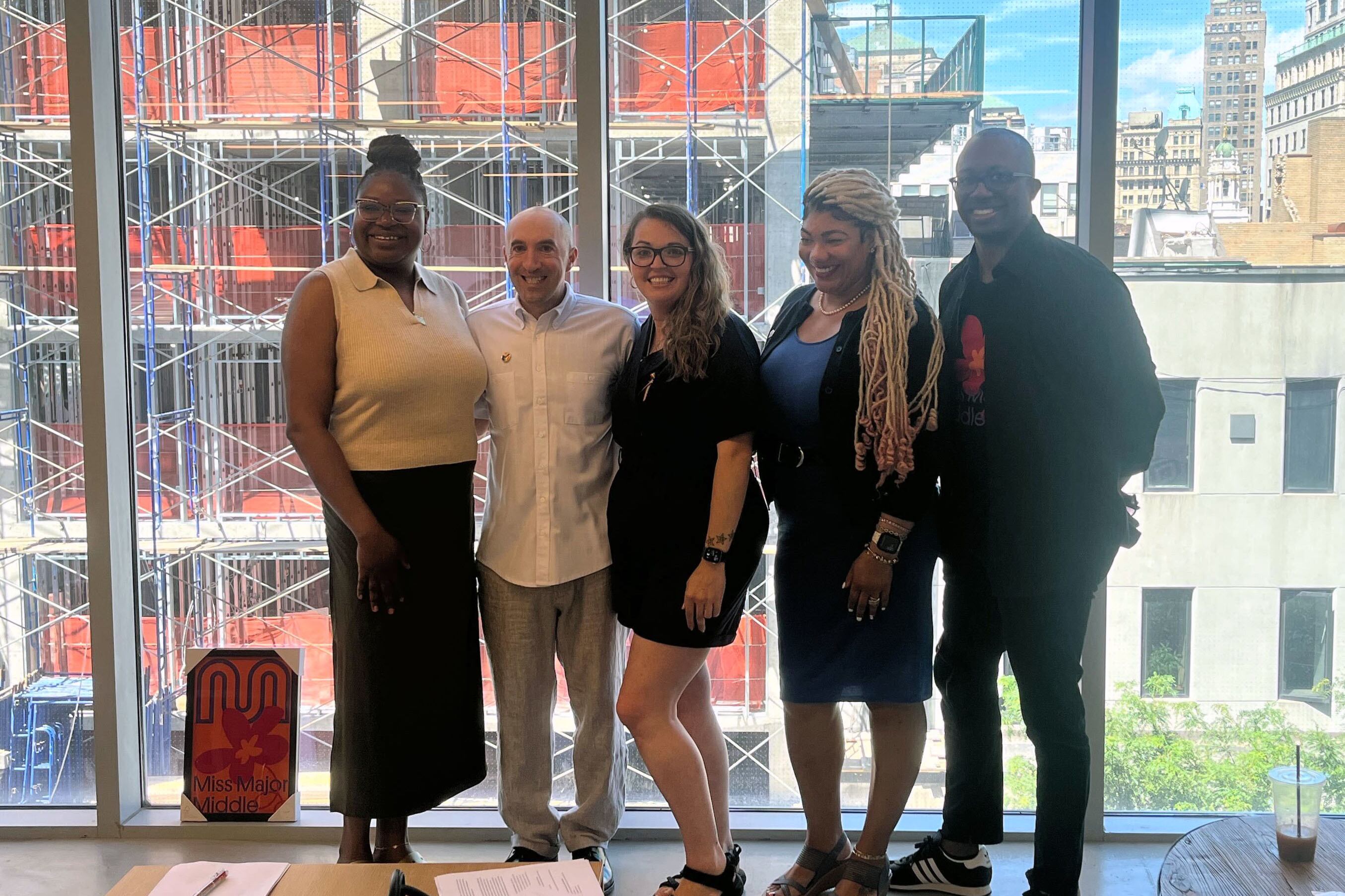 Five people stand side by side posing for a portrait in front of a glass wall.