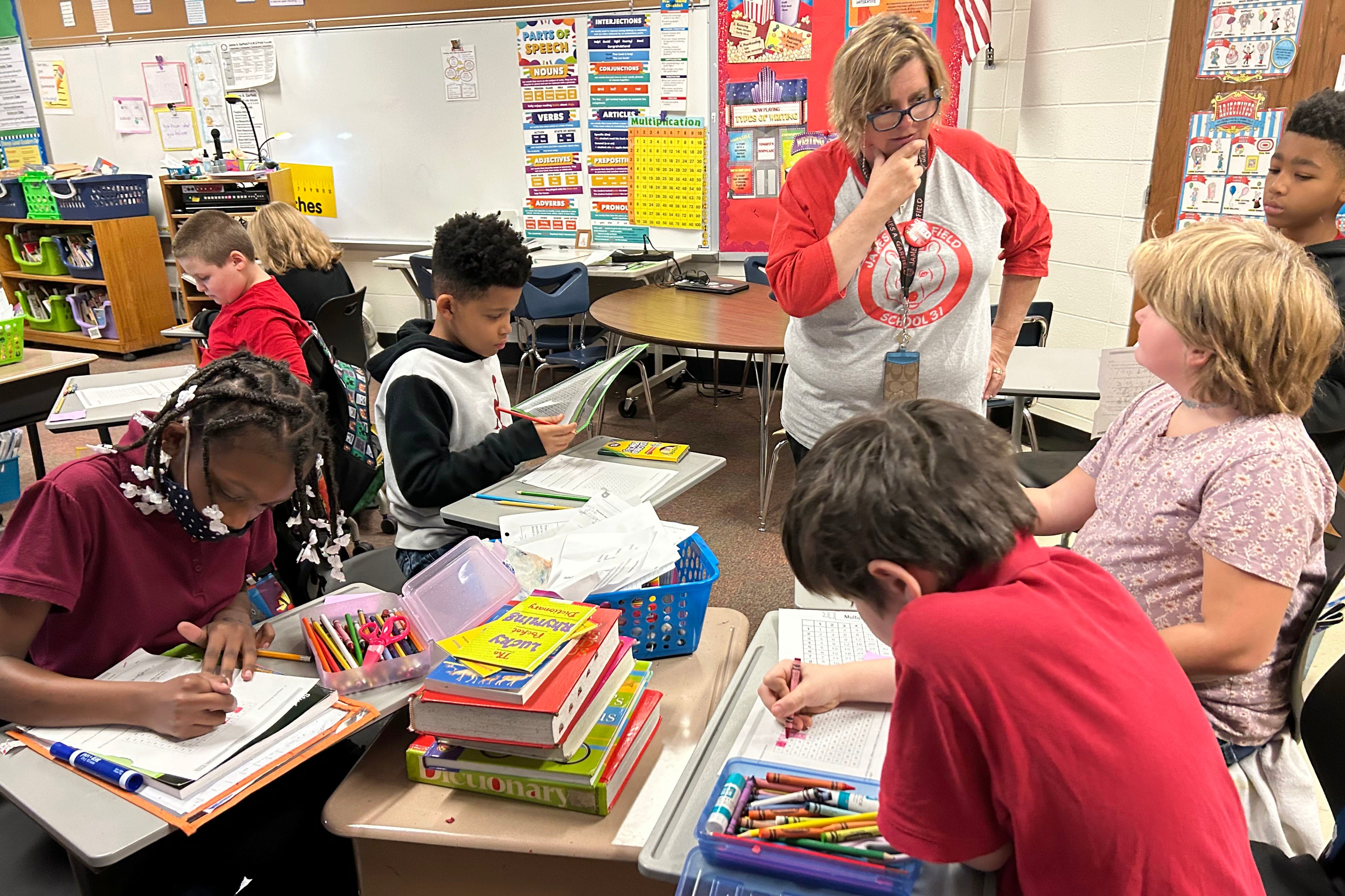 A woman with glasses stands before four students seated at desks doing class work.
