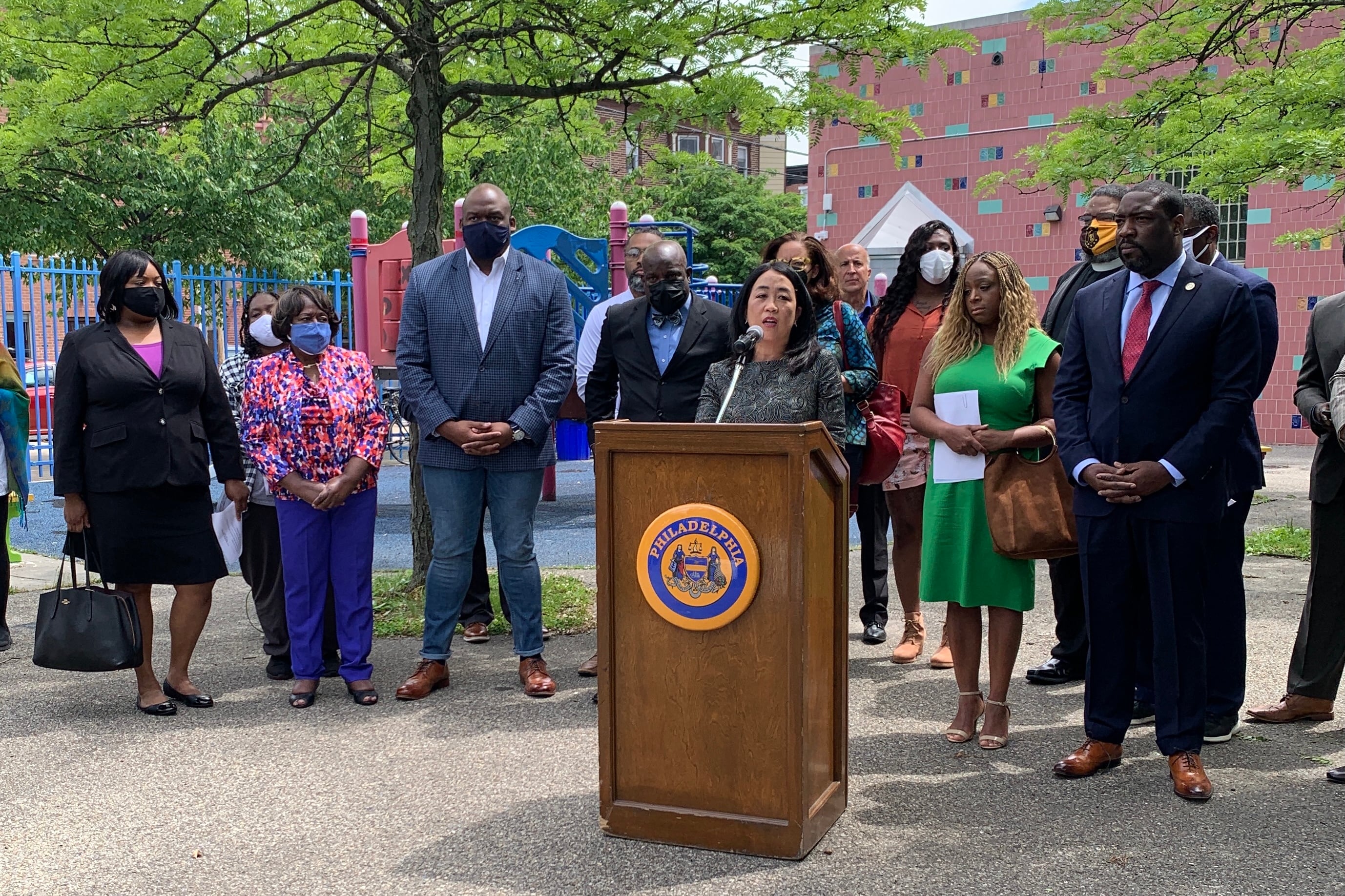 Council member Helen Gym is standing at a lectern outside and is surrounded by several other council members, community and religious leaders.