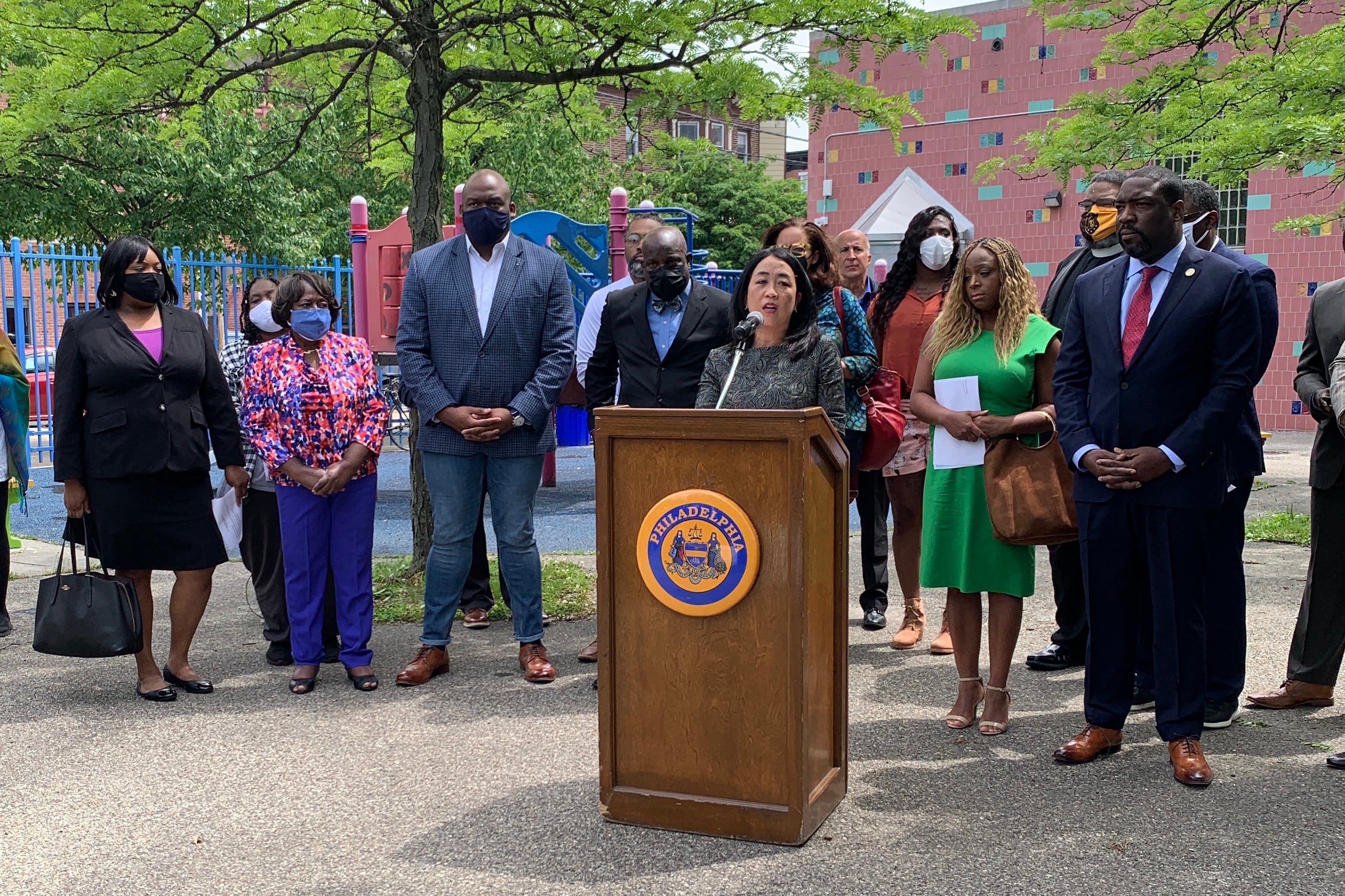 Council member Helen Gym is standing at a lectern outside and is surrounded&nbsp;by several other council members, community and religious leaders.