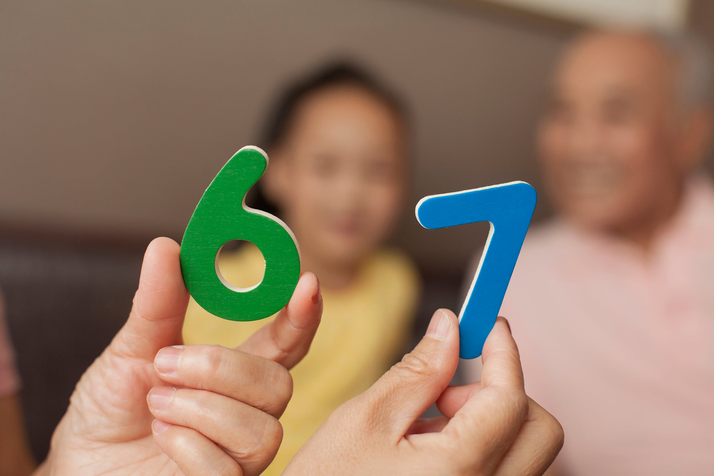 A photograph of a pair of hands holding up number blocks of 6 and 7 with two people sitting in the background.