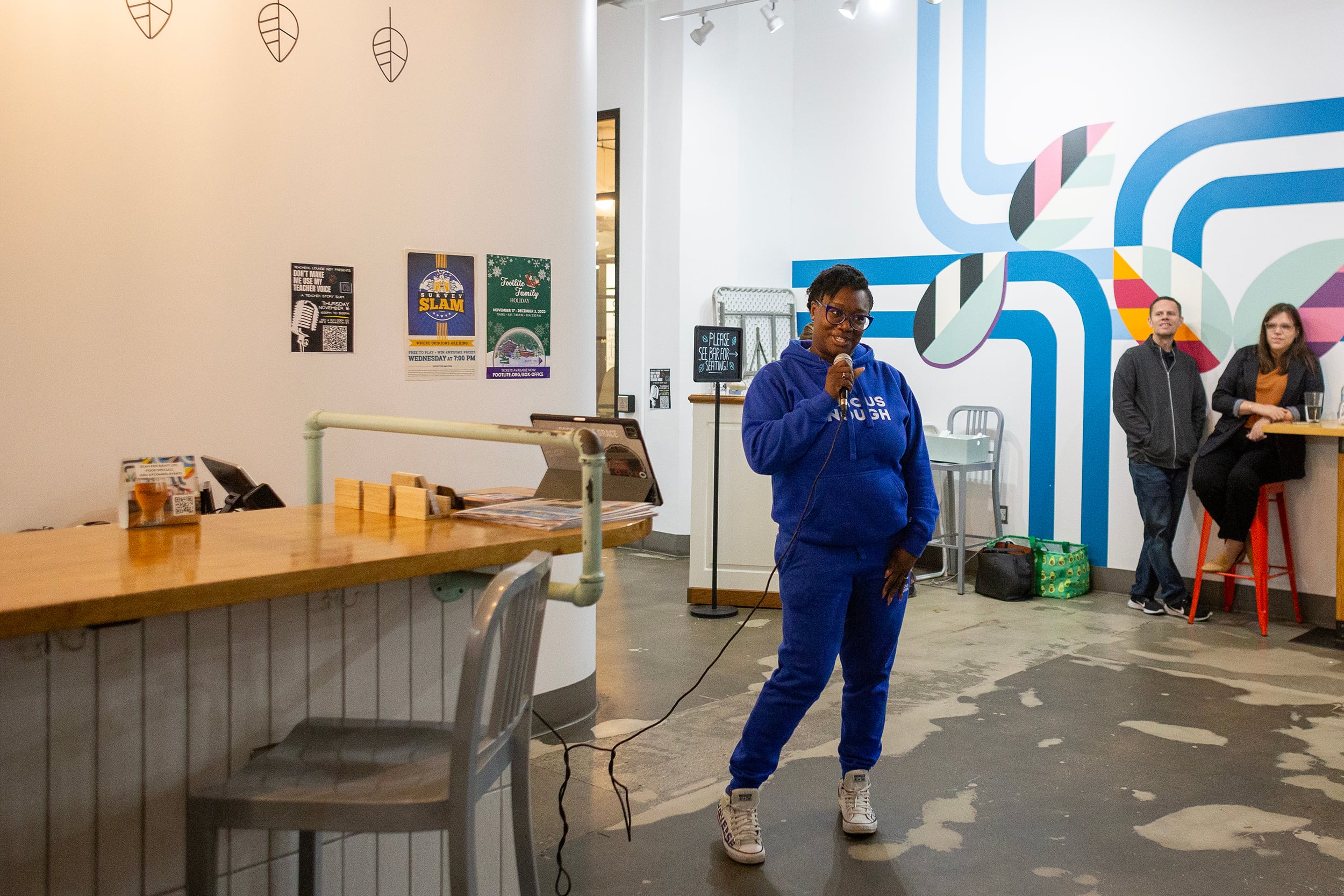 A woman wearing all blue speaks into a microphone while two people sit against the back wall.