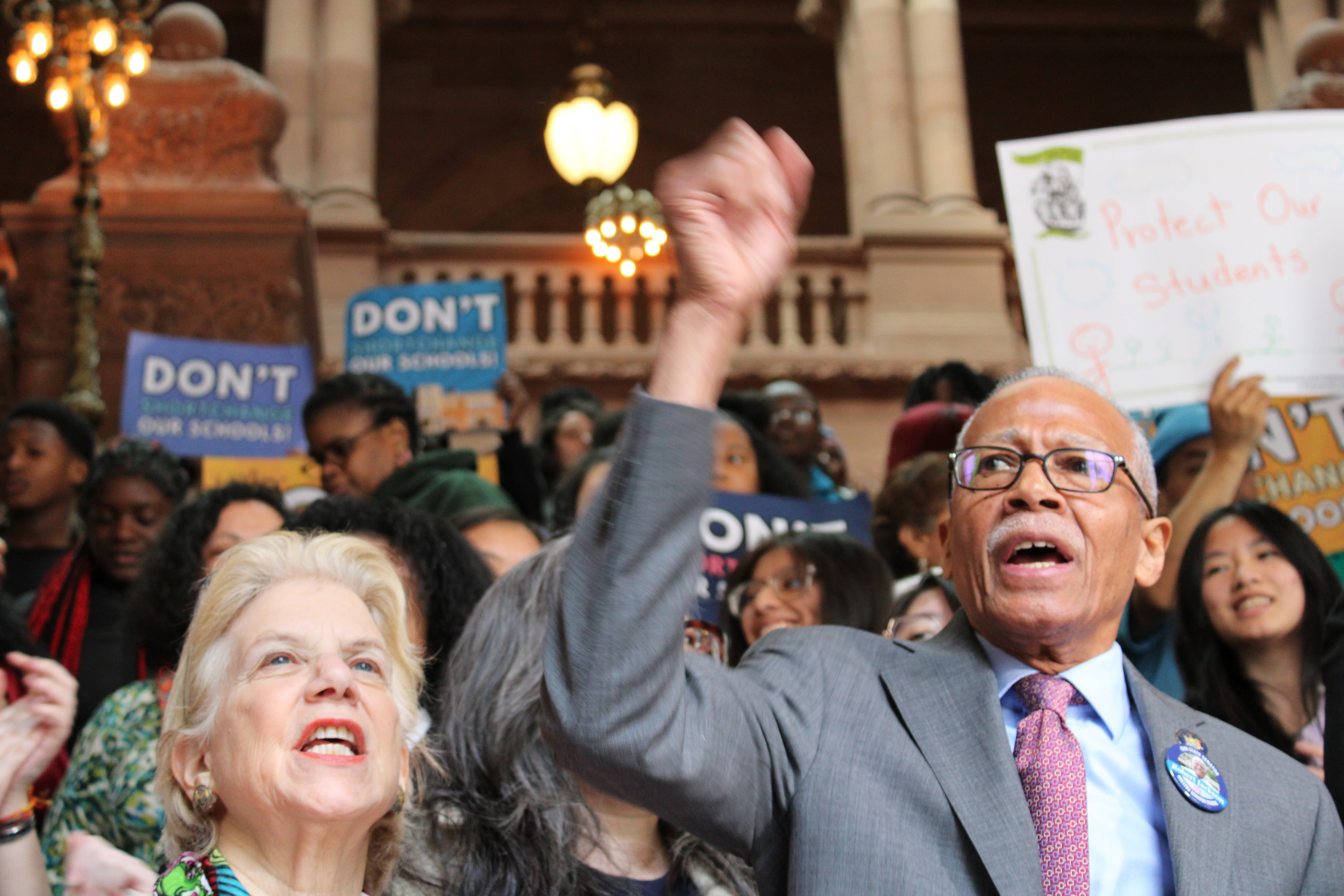 A white woman and a Black man cheer during a protest with a large group of people in the background.