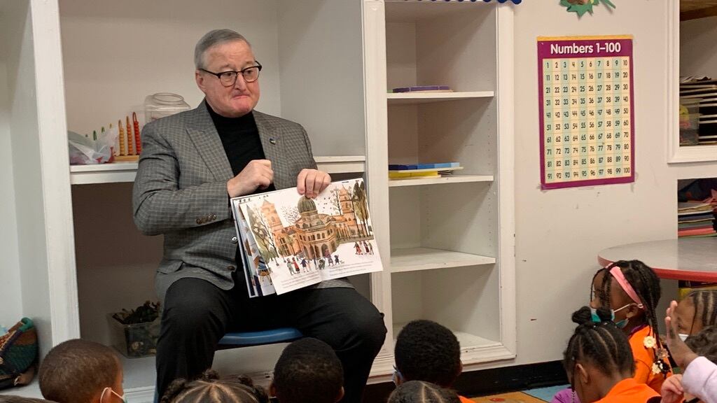 A man wearing glasses and black pants holds a book out for children sitting on the floor and wearing orange shirts to see.