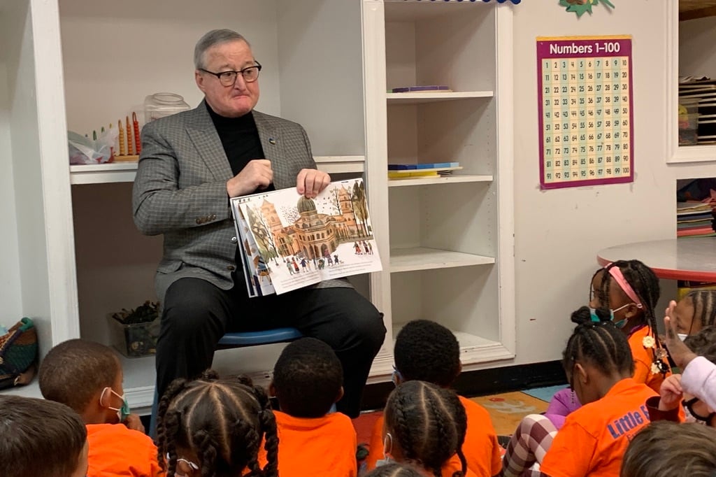 A man wearing glasses and black pants holds a book out for children sitting on the floor and wearing orange shirts to see.