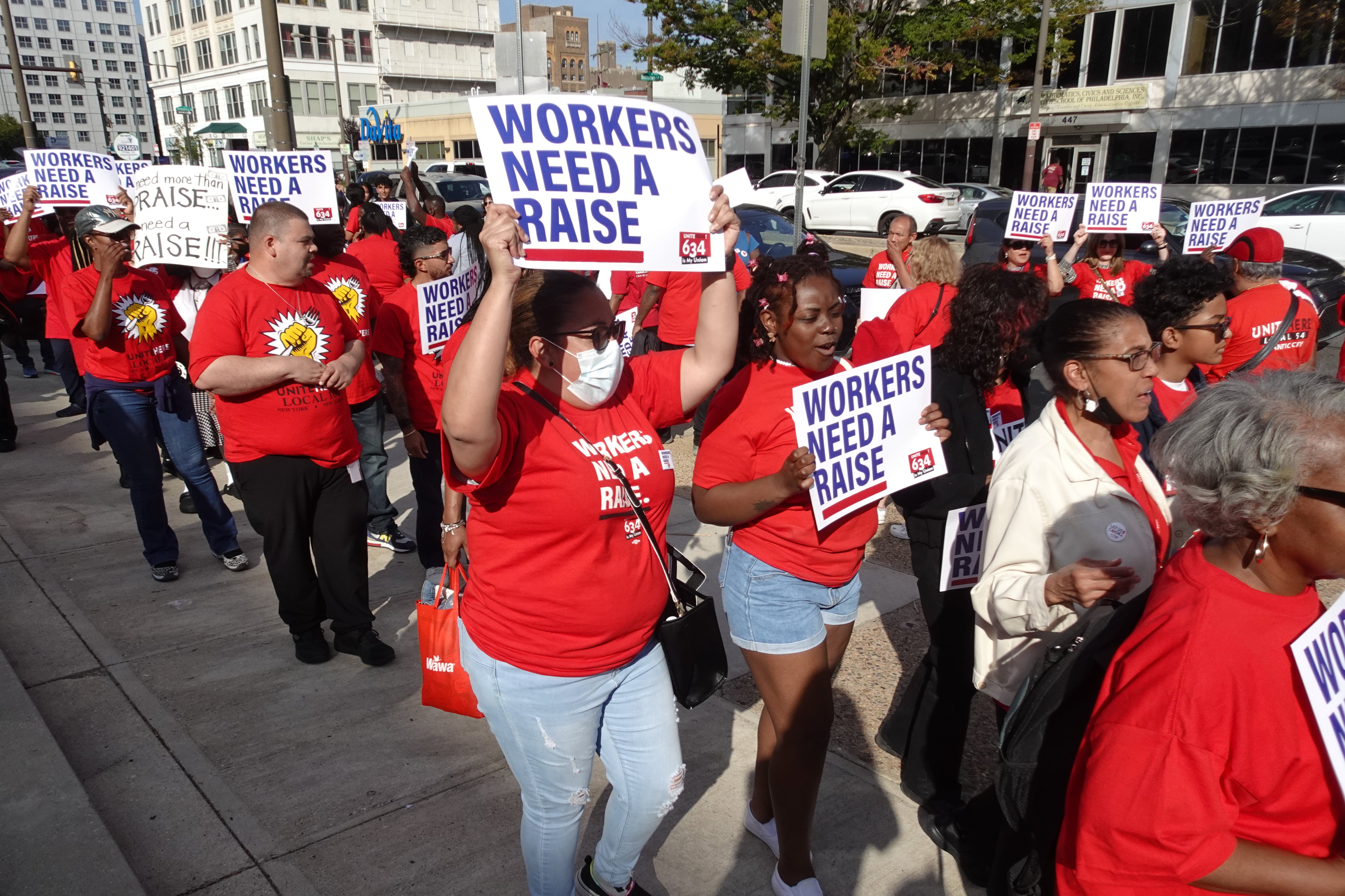 A group of people in red shirts hold signs that say “workers need a raise.”