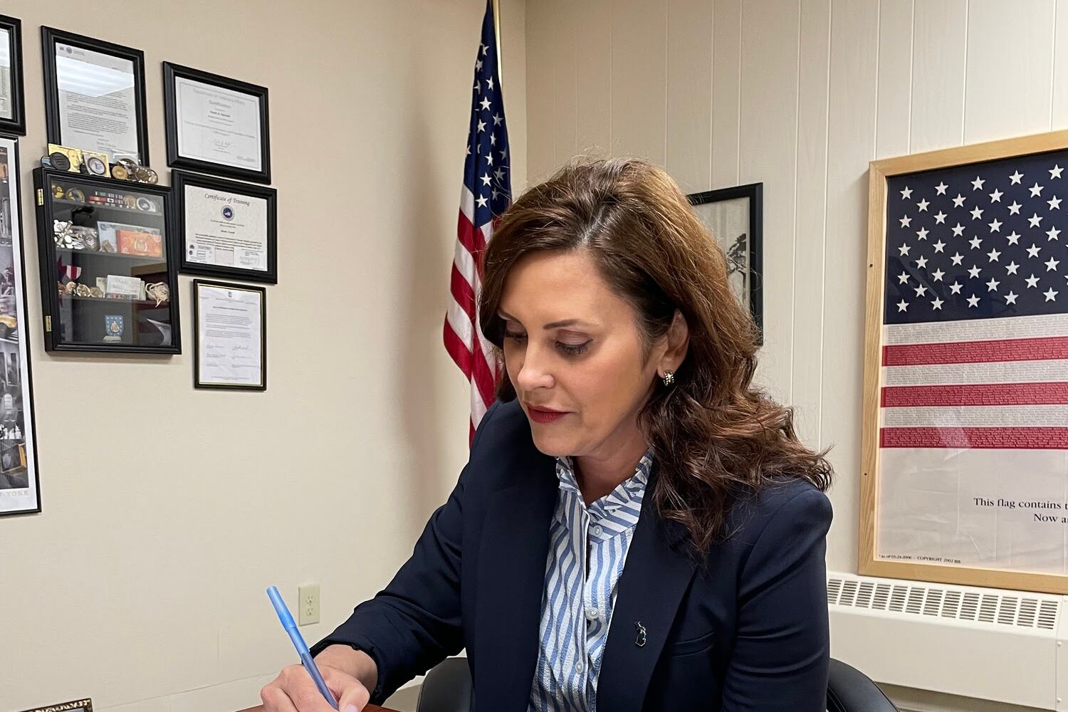 Michigan Governor Gretchen Whitmer signs a bill, sitting at a finished wooden desk in an office.