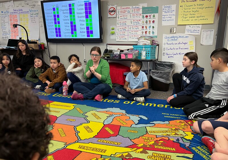 A group of young students and a teacher sit in a classroom with a colorful rug.