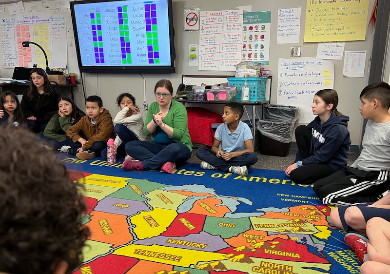 A group of young students and a teacher sit in a classroom with a colorful rug.