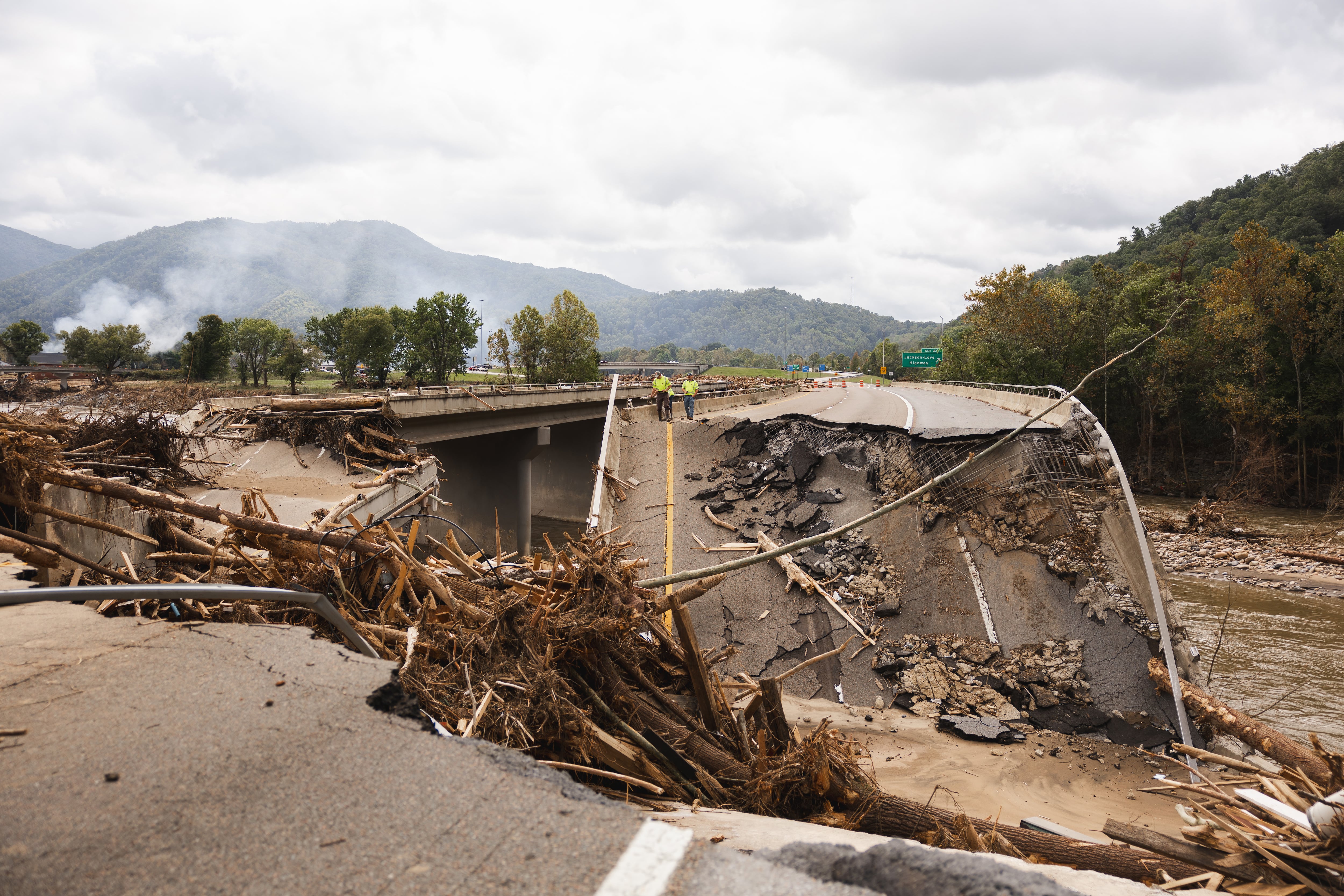 Massive destruction of a bridge and road with a mountain and greenery on the side and background.