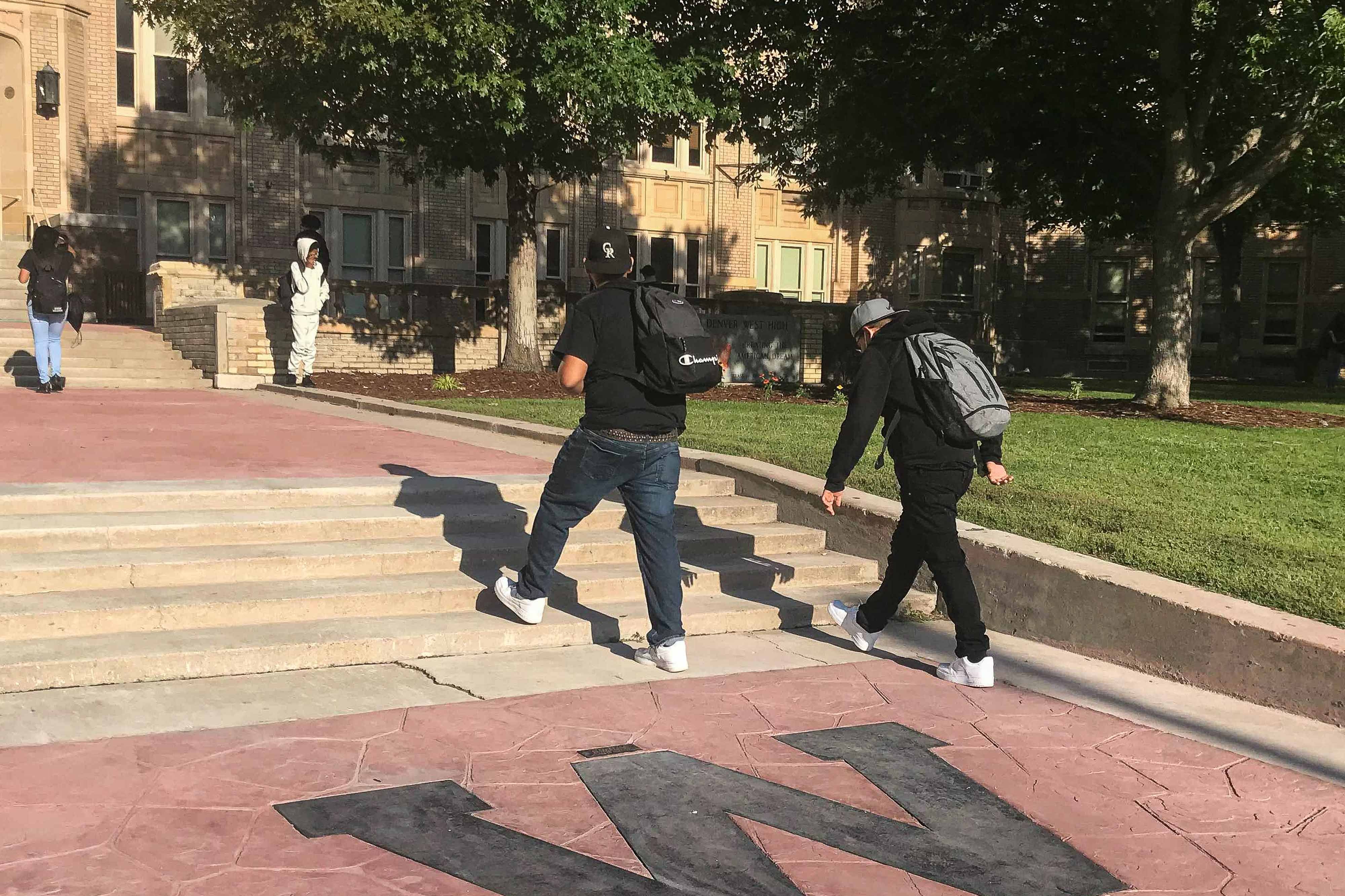 Two high school students wearing black shirts, jeans, and backpacks walk up the front steps of Denver’s West High School, past a black “W” on the red flagstone walkway.