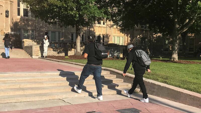 Two high school students wearing black shirts, jeans, and backpacks walk up the front steps of Denver’s West High School, past a black “W” on the red flagstone walkway.