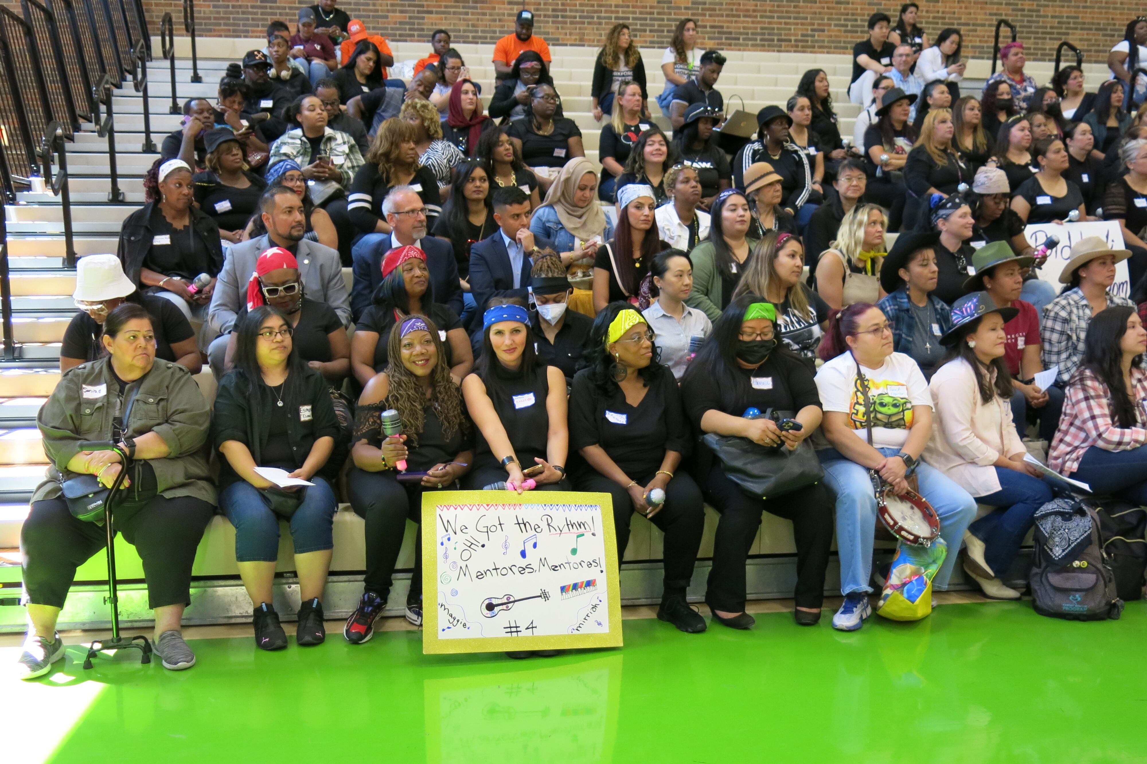 A group of people sit in the bleachers at Harry S Truman Community College in Chicago. Holding signs along a bright green floor.